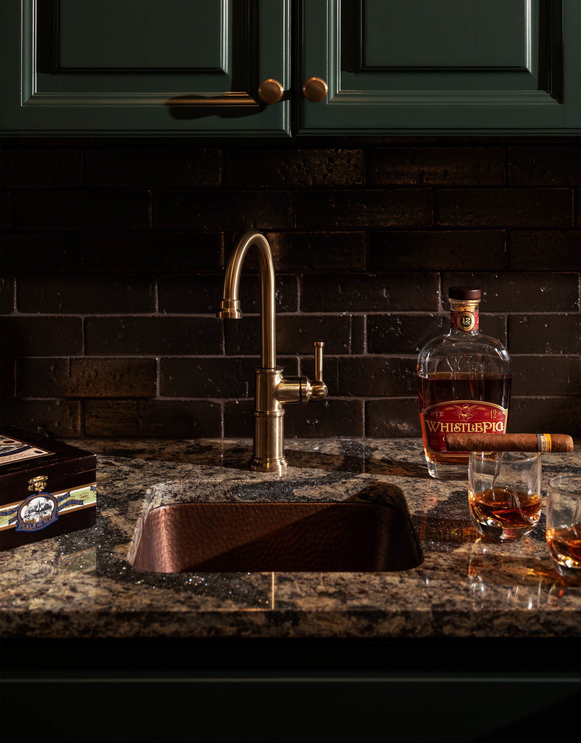 A kitchen countertop with a gold-colored faucet, a bottle of whiskey labeled 'WhistlePig,' a cigar, and three glasses filled with whiskey, with dark green cabinets above and a dark brick backsplash.