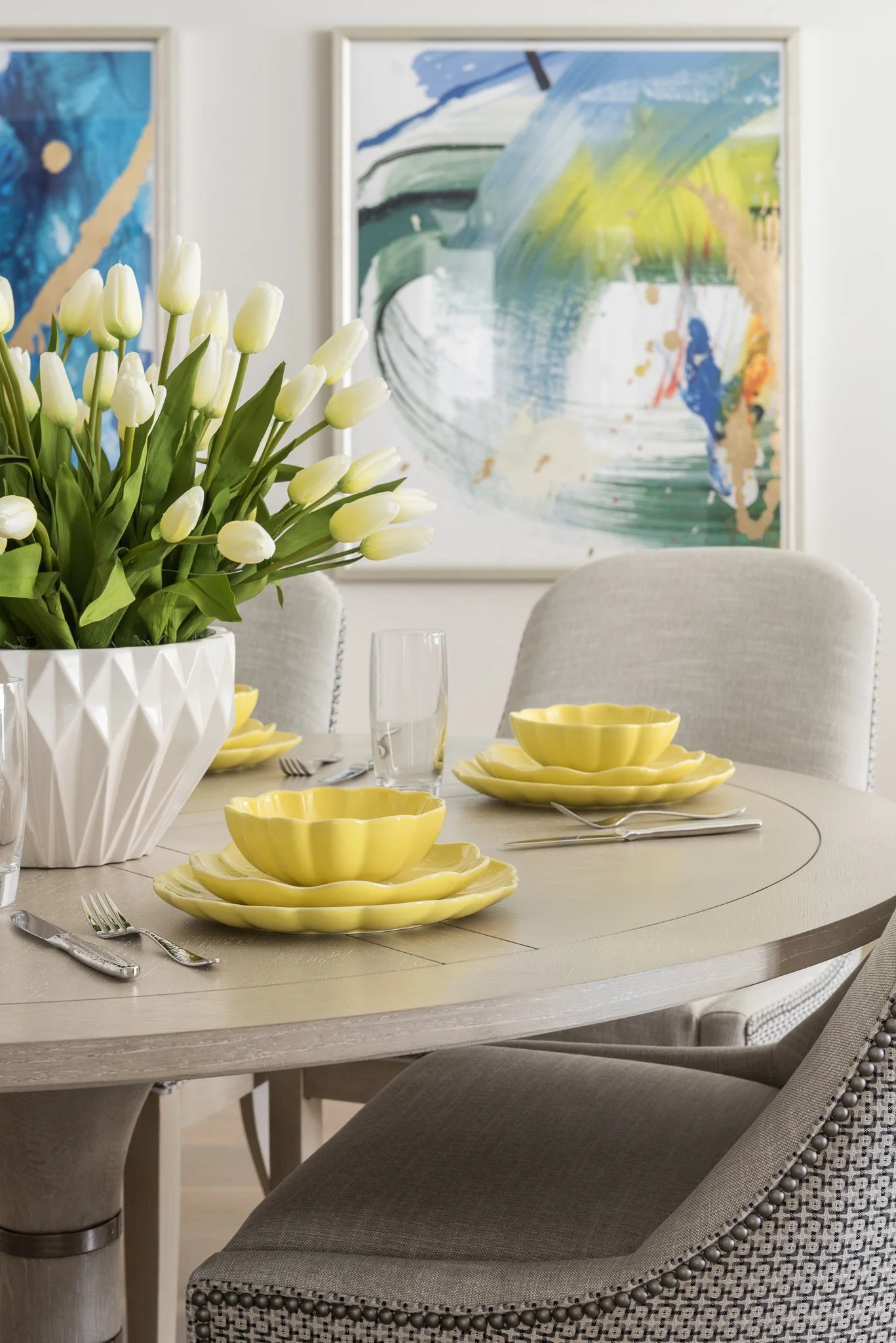 A dining room table set with yellow scalloped dishes, silverware, and clear glasses, decorated with a large white vase of white tulips, with modern abstract art on the wall in the background.
