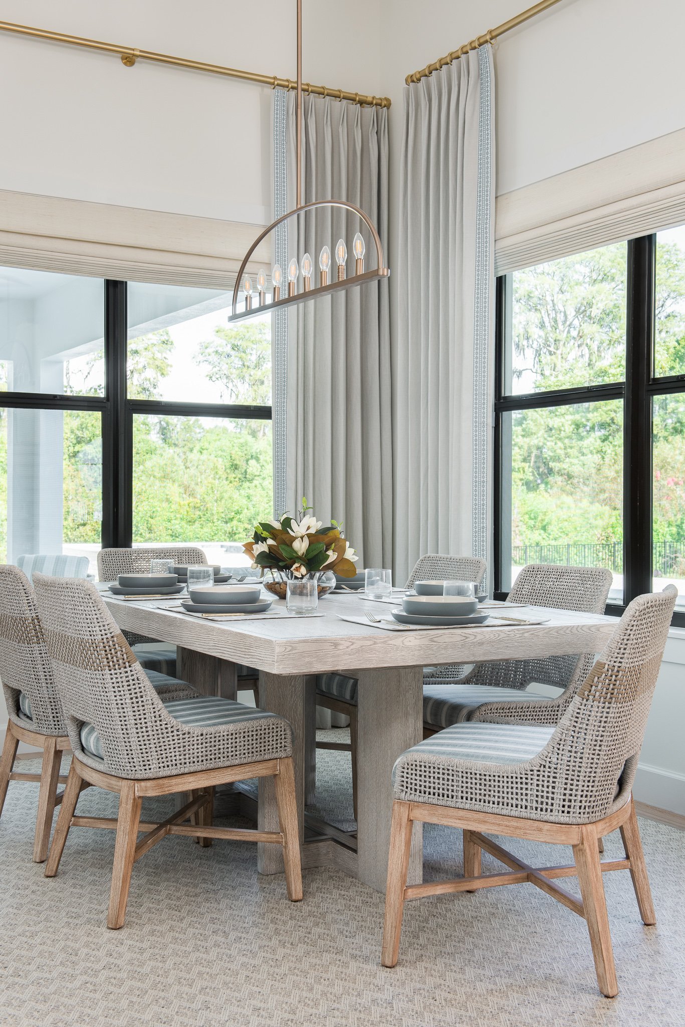 A dining room with a wooden rectangular table set with white dishes, glasses, and a flower centerpiece. There are eight woven chairs with striped cushions around the table. The room has large windows with black frames, cream-colored curtains, and a m