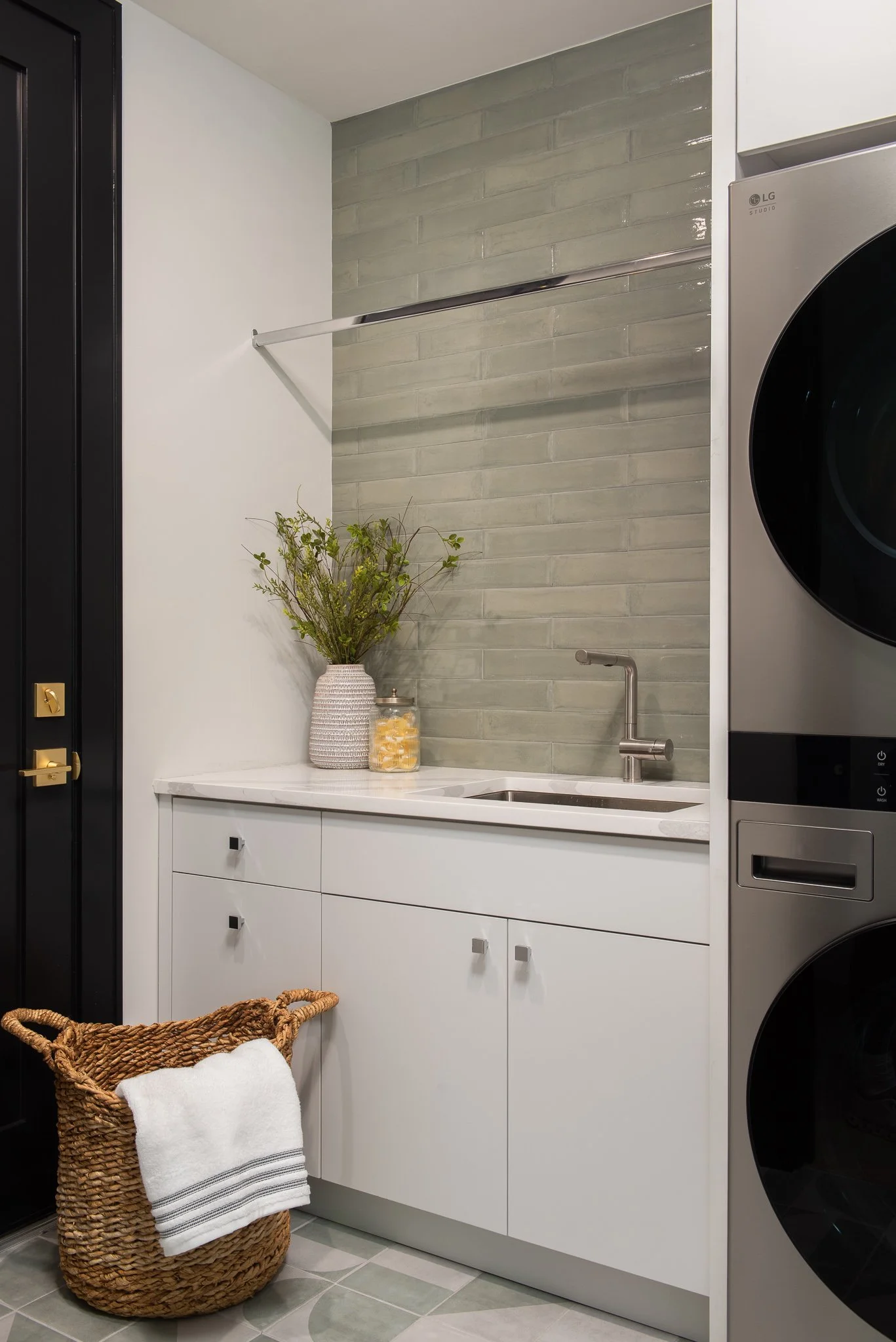 Laundry room with white cabinets, a green tiled wall, a washing machine and dryer, a small sink, a wicker basket with a white towel, decorative vases with greenery, and a black door.