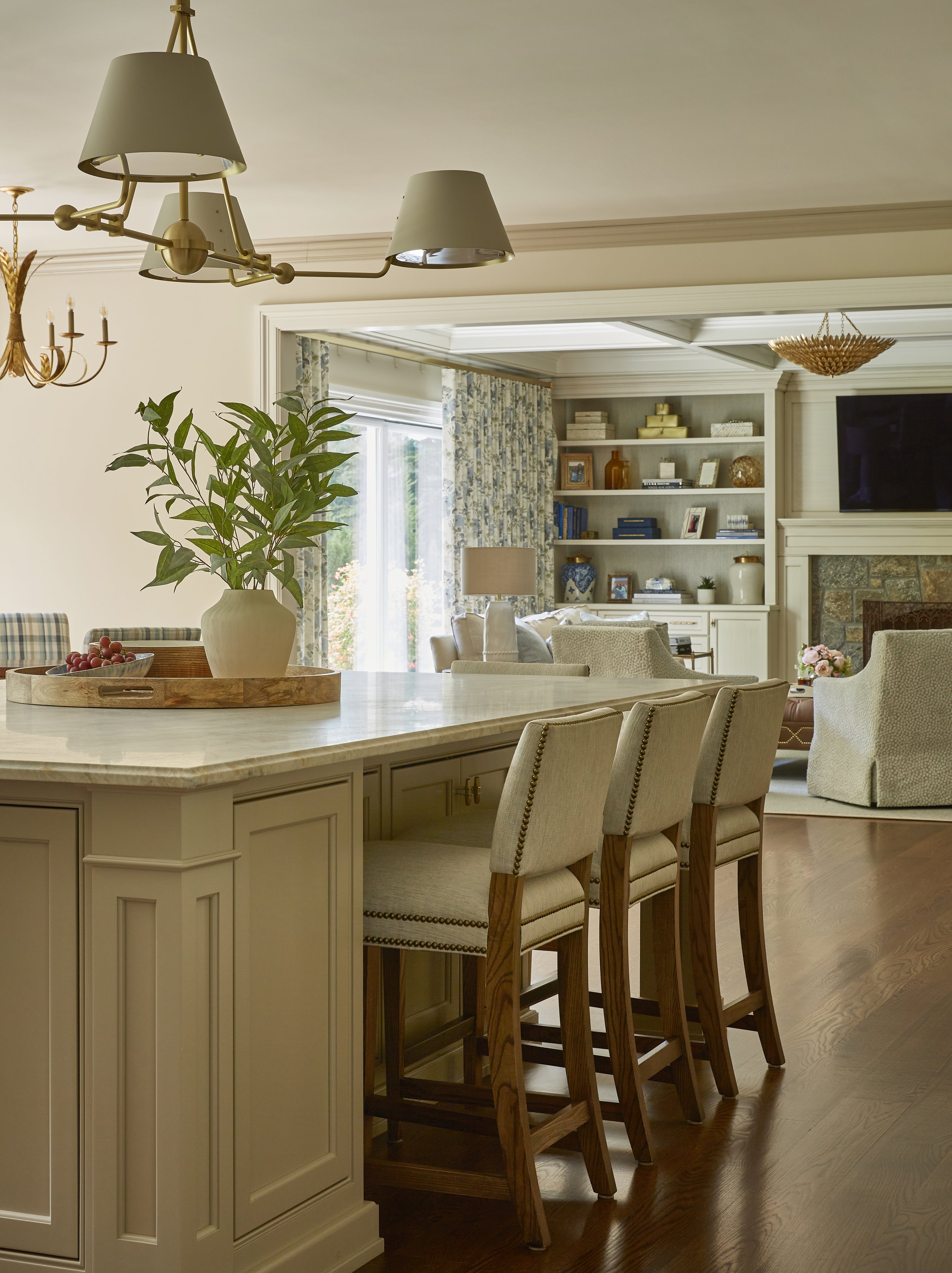 A bright, cozy living space with a kitchen island, four barstools, and a view into a living room with bookshelves, a fireplace, and a large flat-screen TV.