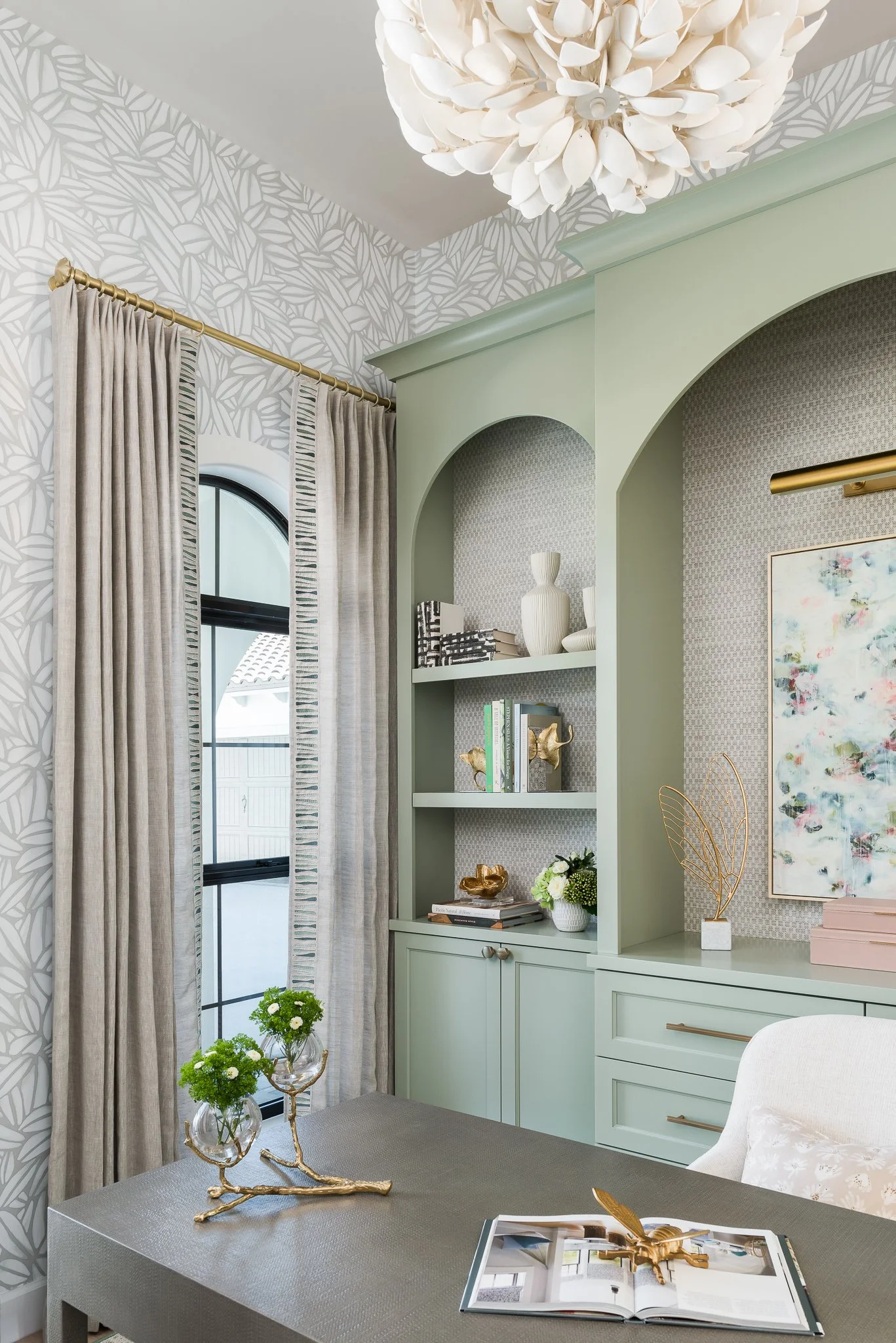 Interior of a room with light green built-in shelves, a window with beige curtains, a light-colored patterned wallpaper, a white chair, and a chandelier overhead. The table has a decorative gold branch with small green plants, and an open magazine.