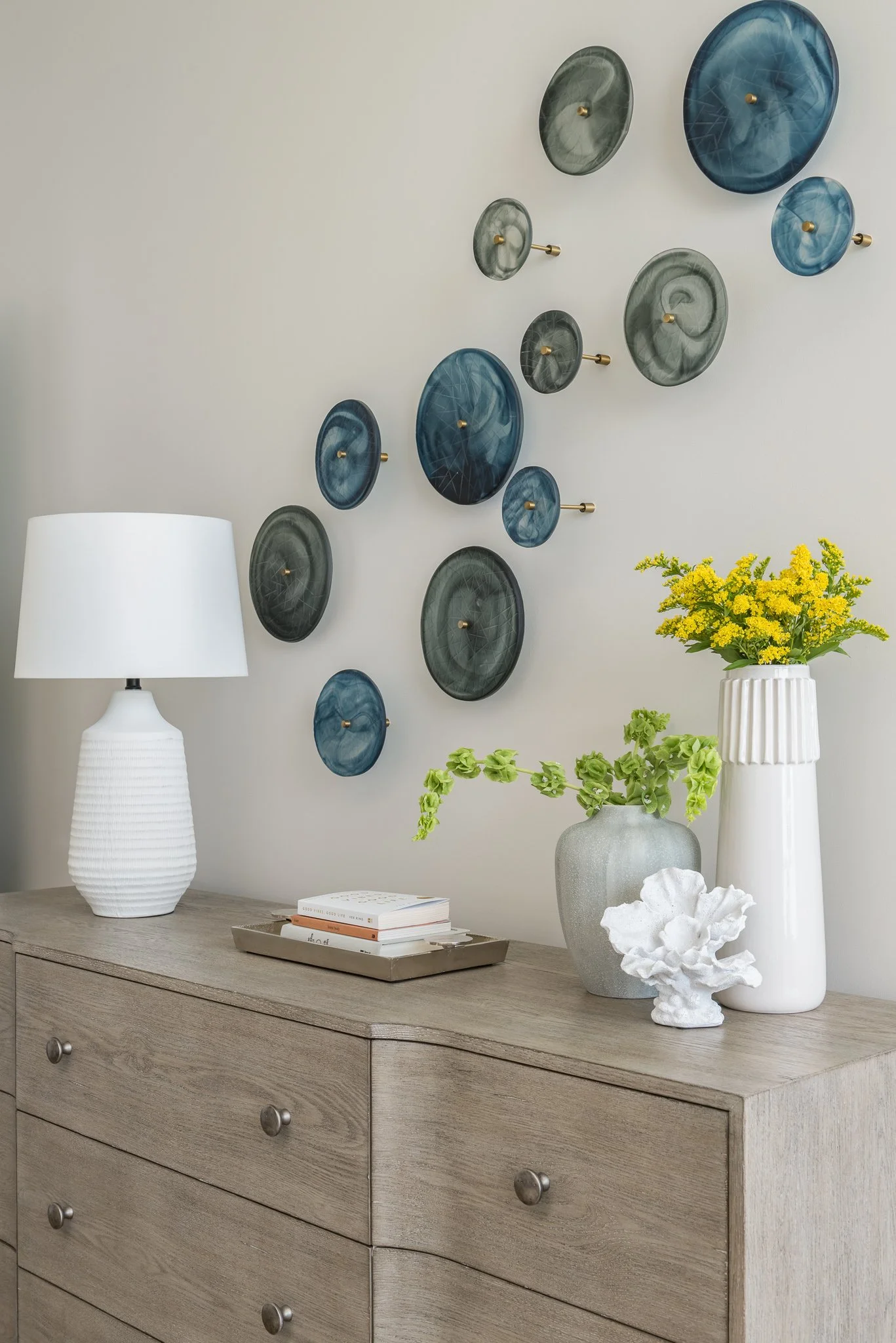 Decorative wall art featuring various circular plates in shades of blue and gray arranged above a wooden dresser with a white table lamp, books, and vases with yellow flowers and green foliage.