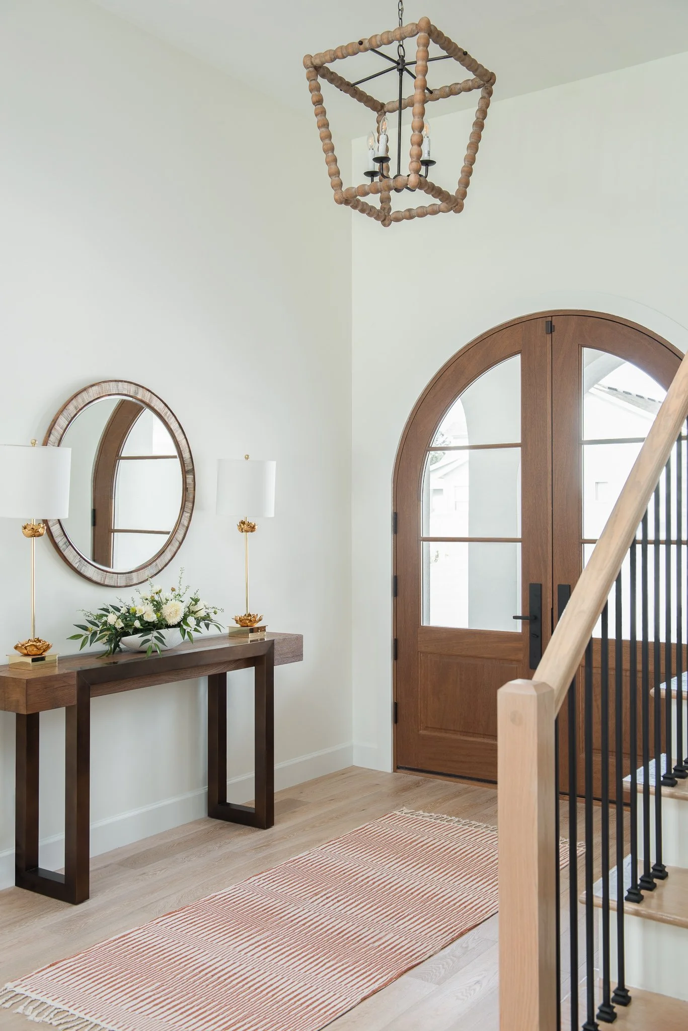 Entryway with wooden door, a long wooden console table with a floral arrangement and two white lamps, a round wall mirror, a striped rug, a staircase with black metal railing, and a geometric wooden and metal chandelier hanging from the ceiling.
