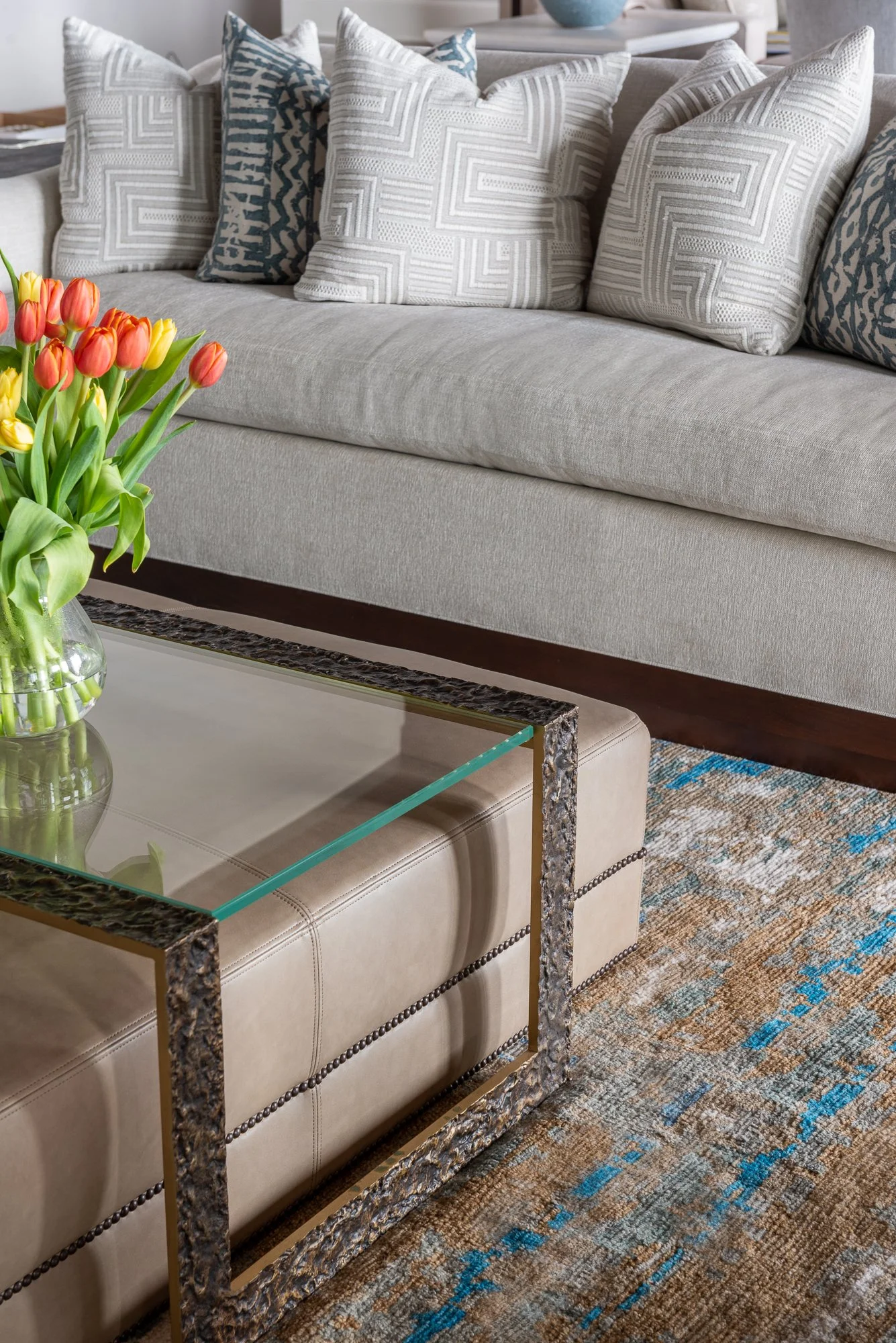 A living room with a light gray sofa decorated with white and blue patterned cushions, a glass-topped coffee table with a beige upholstered base, and a glass vase filled with orange and yellow tulips on the table. There is a multicolored area rug wit