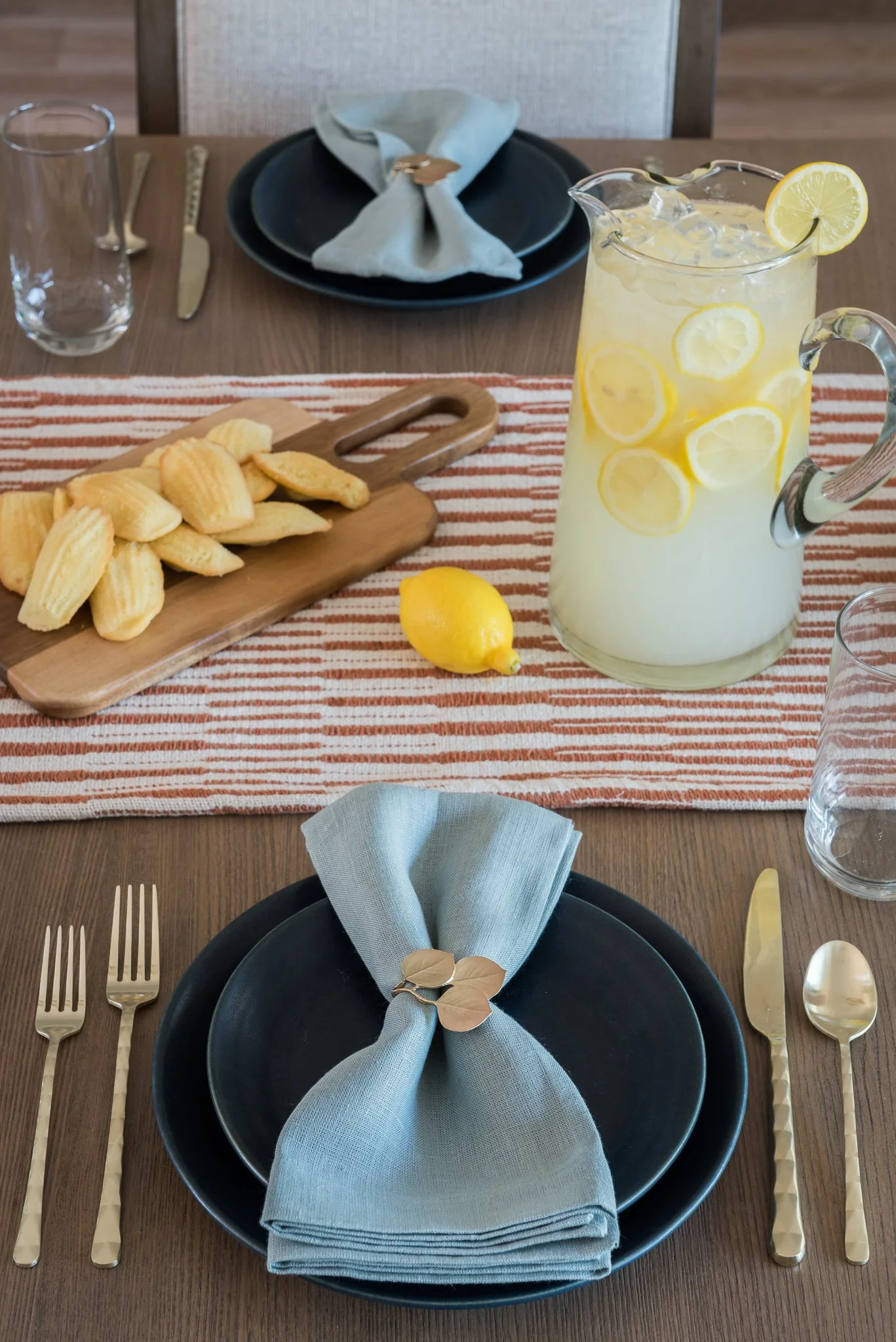 A dining table set for two with black plates, gold utensils, blue napkins, a striped table runner, a wooden serving board with madeleines, a pitcher of lemonade with lemon slices, and a whole lemon, with a gray upholstered chair in the background.
