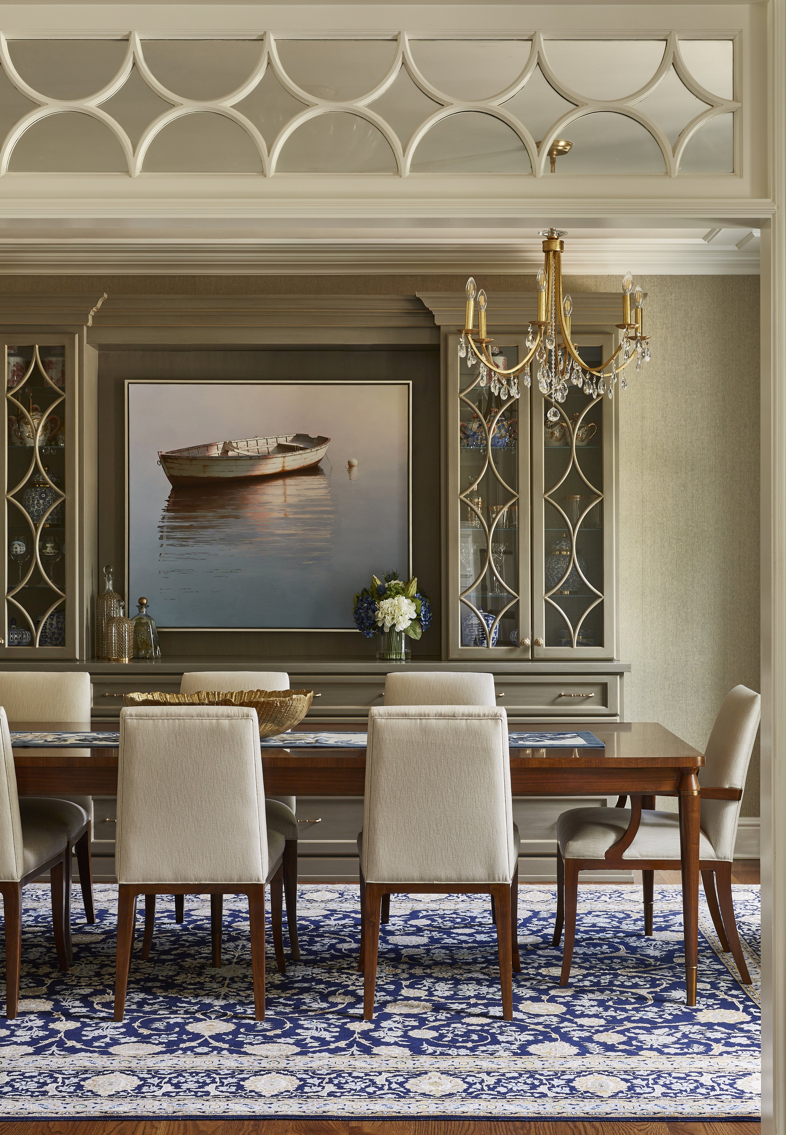 Elegant dining room with a wooden table and six upholstered chairs, a chandelier hanging above, a large framed photo of a boat on water, glass cabinets on either side with decorative items, a vase with flowers on the cabinet, and a patterned blue and