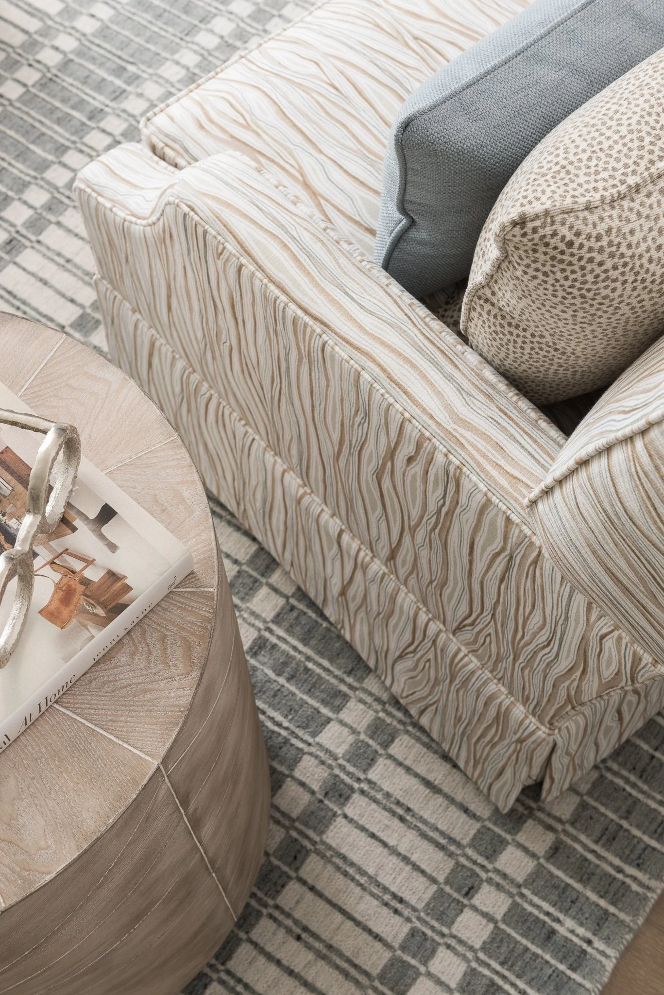 Close-up of a living room corner showing a beige and cream striped upholstered sofa with blue and beige patterned pillows, a round wooden coffee table with a magazine and decorative object, and a patterned area rug.
