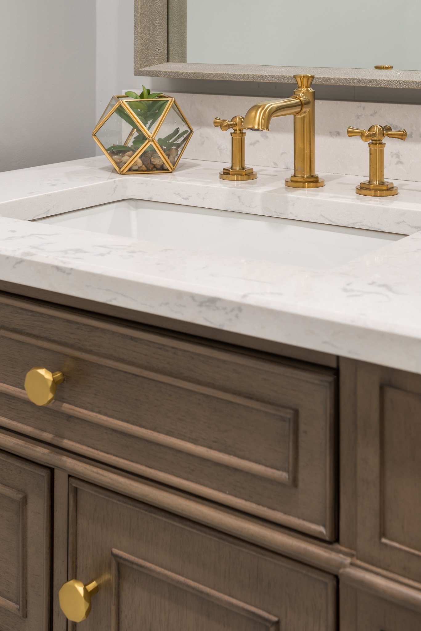 A bathroom vanity with a marble countertop, gold fixtures, and a decorative glass and gold plant holder with plants. A mirror is partially visible above.