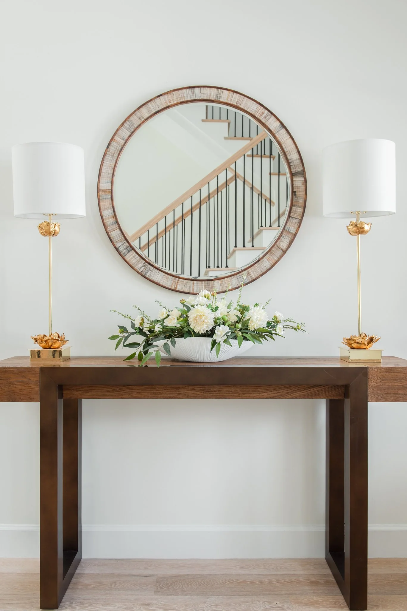 A wooden console table with a floral arrangement in a white vase, flanked by two white lamps with gold accents, against a white wall with a round mirror reflecting stairs.