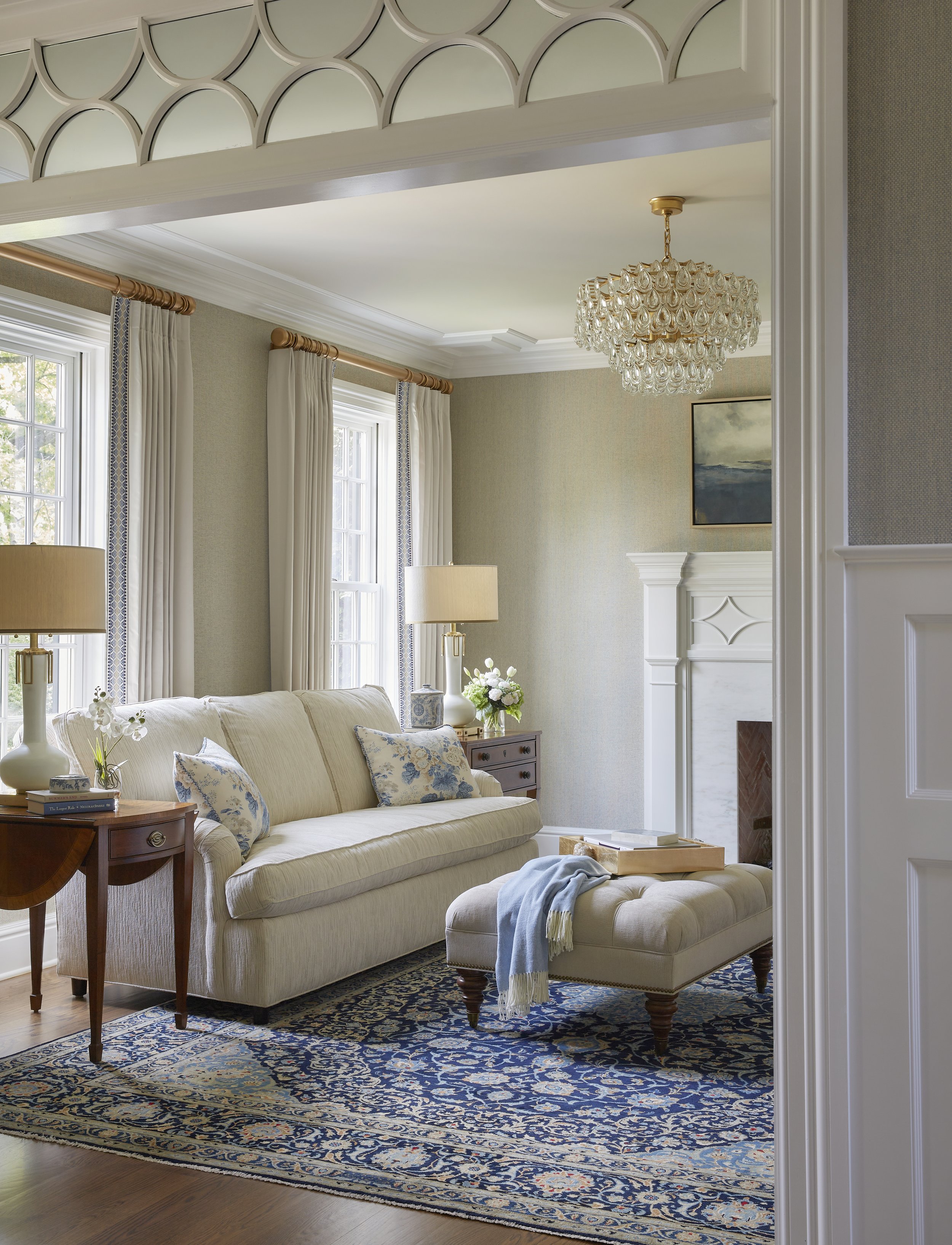 Living room with cream sofa, wooden side tables, blue and cream pillows, beige ottoman with a blue throw, patterned carpet, fireplace, large windows with curtains, chandelier, painting on wall, lamps, flowers, and books.