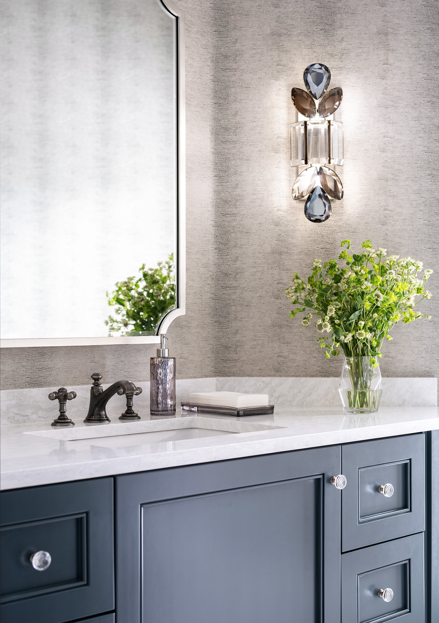 Bathroom vanity with navy cabinets, white marble countertop, and a large mirror. Decorative wall sconce with jewel-like elements, a soap dispenser, and a vase of white flowers are on the counter.