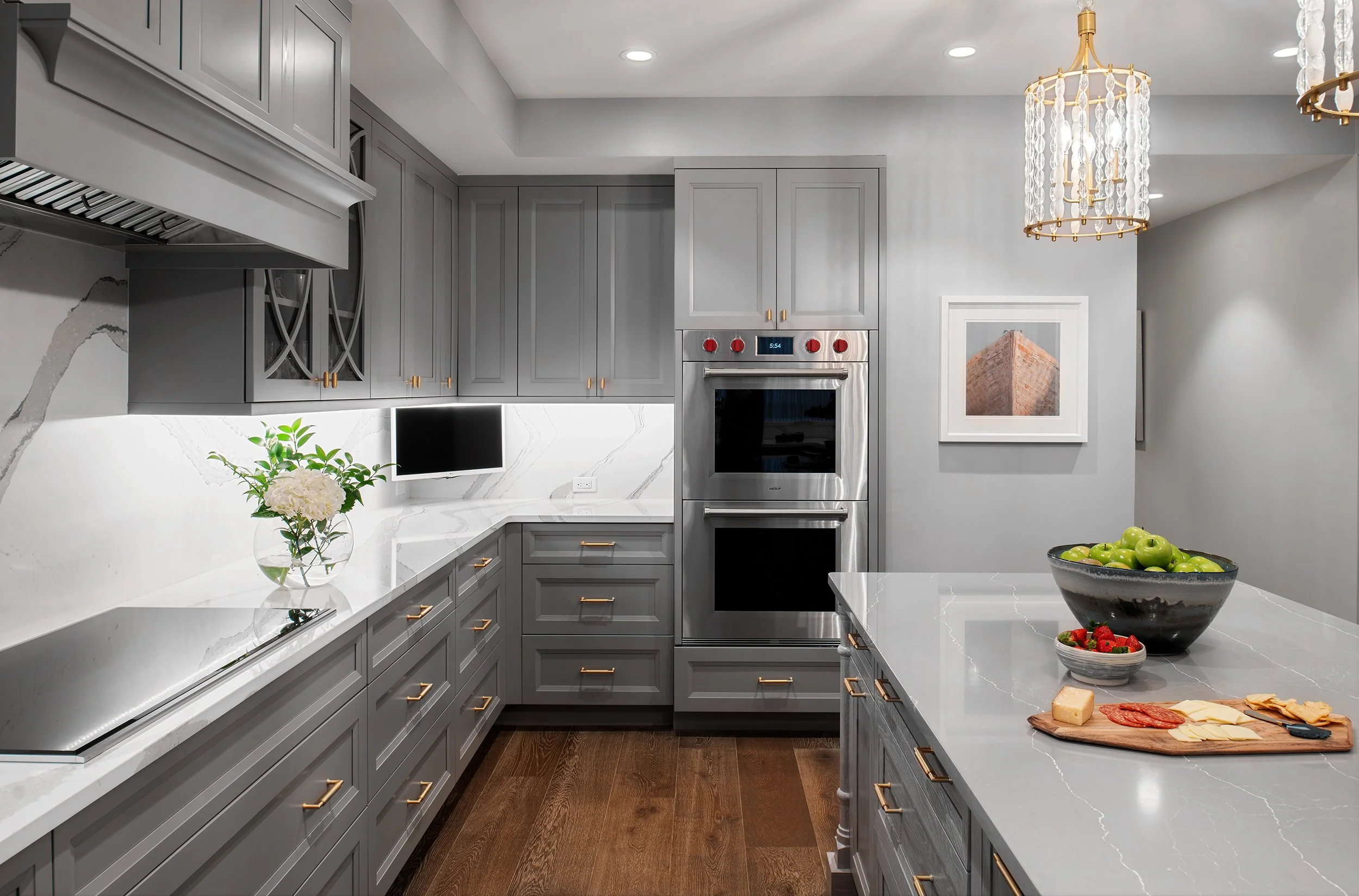 Modern kitchen with gray cabinets, white marble countertops, stainless steel double oven, small wall-mounted TV, decorative art, pendant lights, and a kitchen island with green apples, strawberries, cheese, and meats.