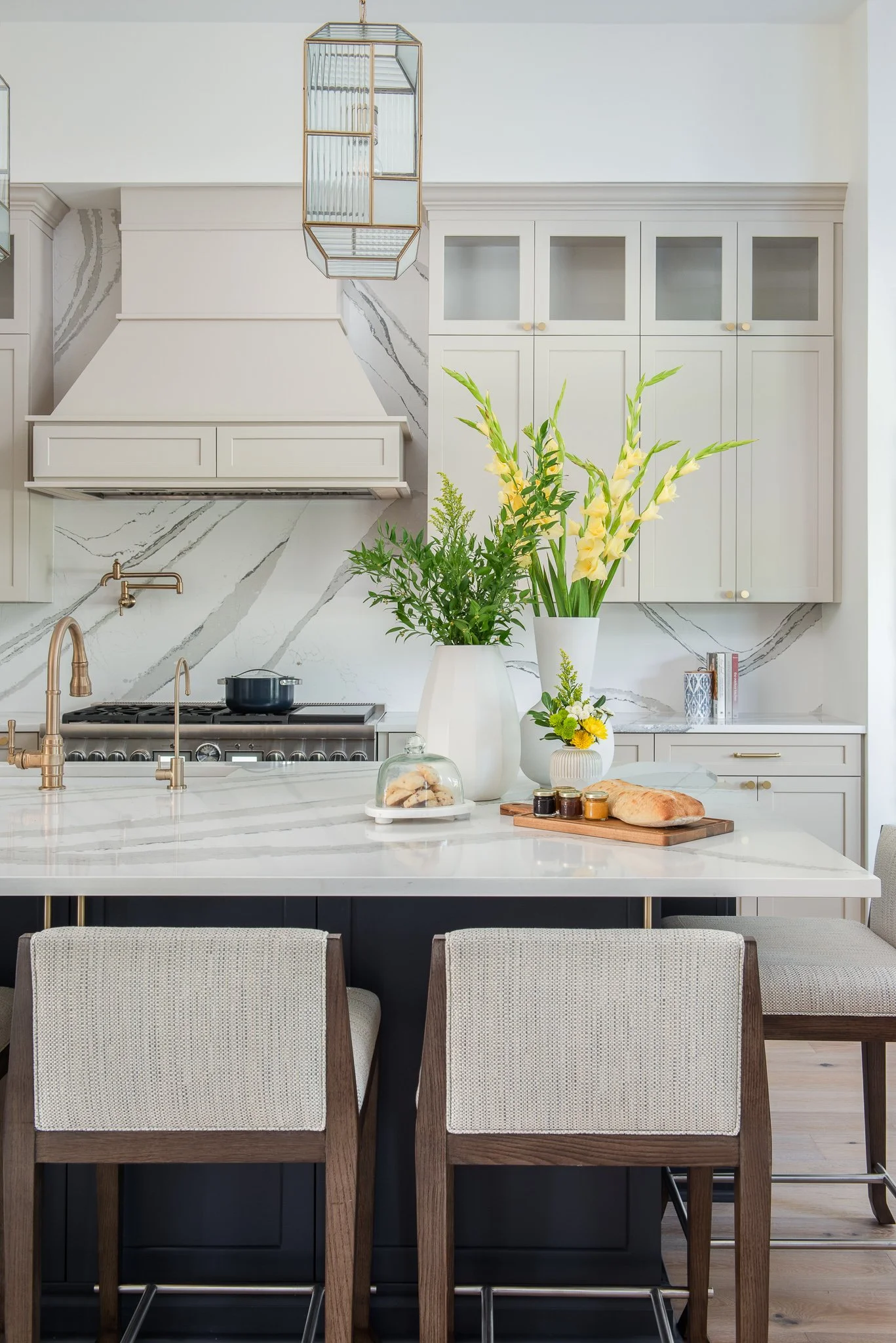 Modern kitchen with white cabinetry, marble backsplash, and a large island with a white countertop. Decor includes tall vases with yellow flowers and green foliage, a loaf of bread, jars, and a cake on a stand. There are three chairs with wooden fram