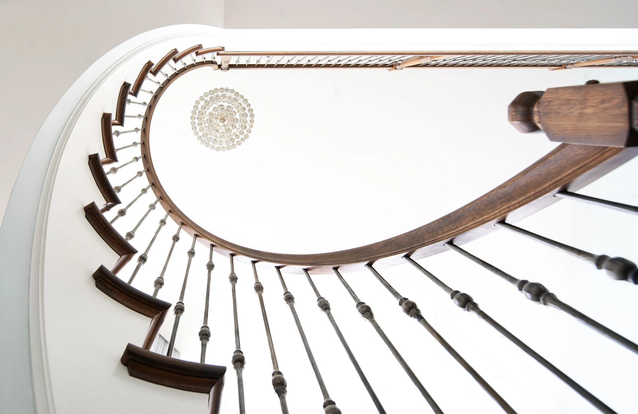 Interior view looking up a spiral staircase with wooden handrail and metal spindles, leading to a chandelier hanging from the ceiling.