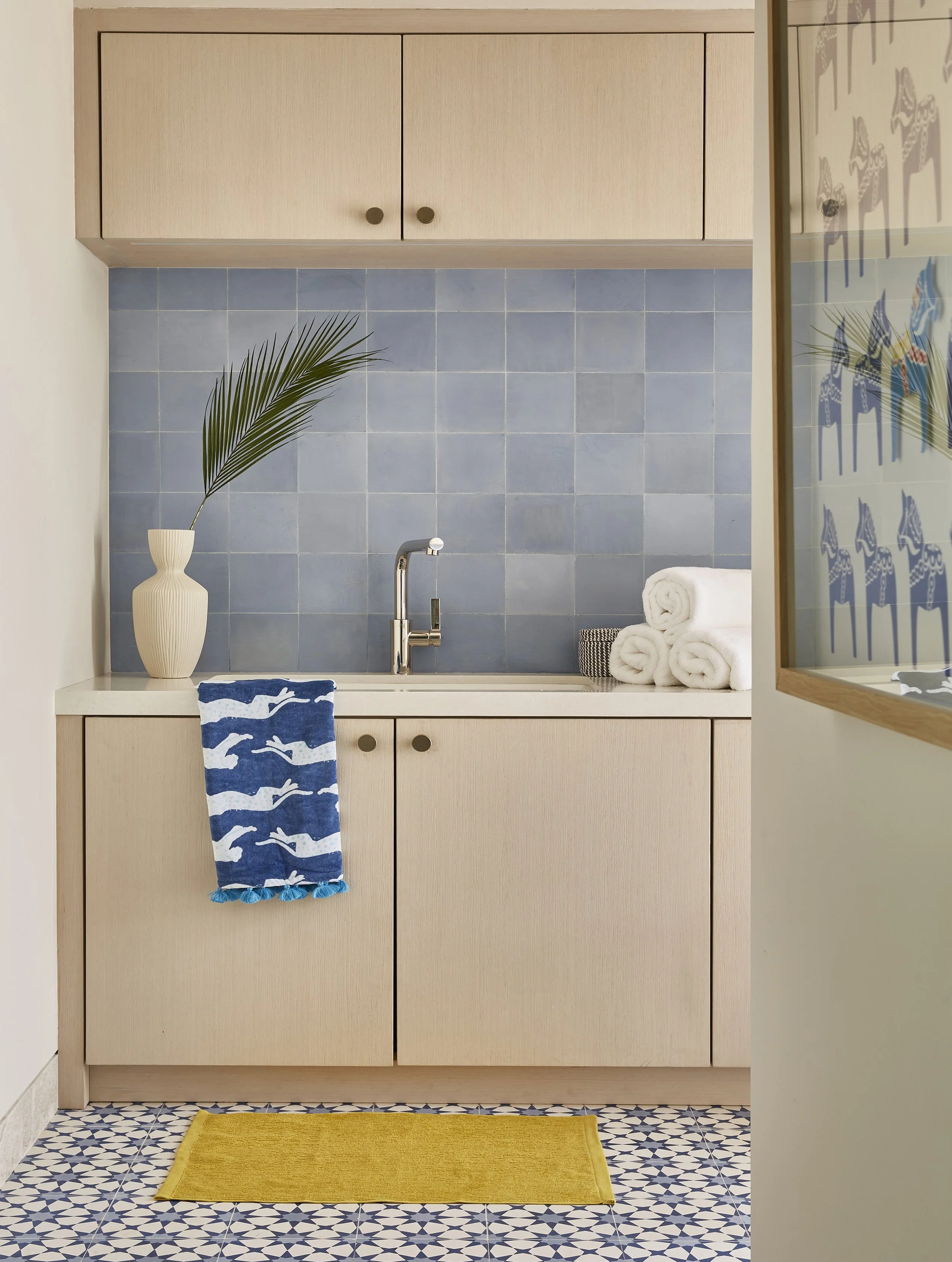 A bathroom countertop with a tiled blue backsplash, a faucet, a striped towel hanging from the cabinet, a white vase with a palm leaf, neatly rolled white towels, and a yellow rug on a blue and white patterned floor.