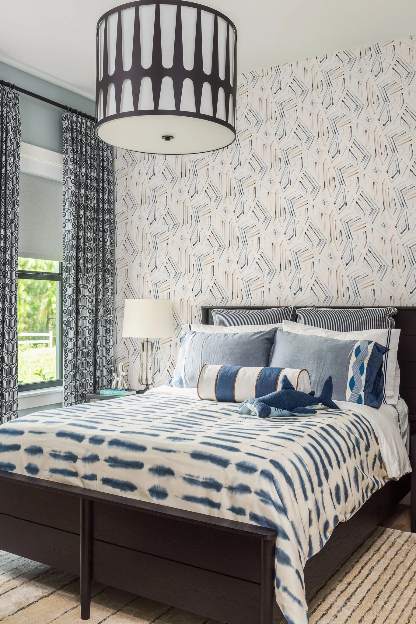 A bedroom with patterned wallpaper, blue and white bedding, a dark wooden bed frame, and a bedside table with a lamp, curtains, and a window showing green outdoors.