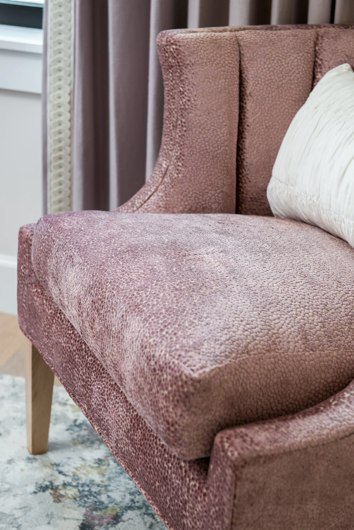 Close-up of a pink textured upholstered armchair with a white pillow on it, in a living room with curtains and a window in the background.