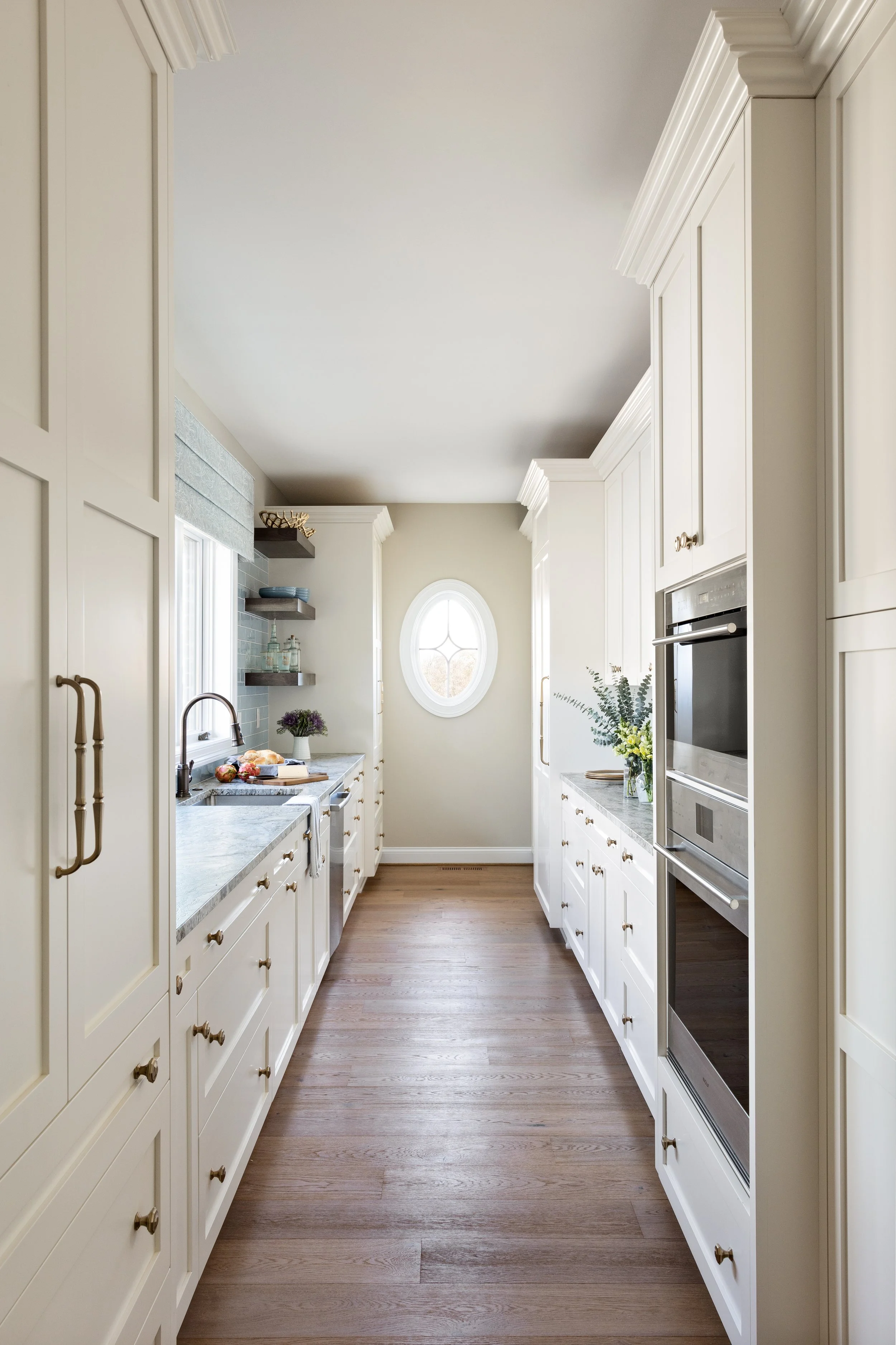 Clean and bright kitchen with white cabinetry, stainless steel appliances, and wooden flooring, featuring a small round window at the back wall.