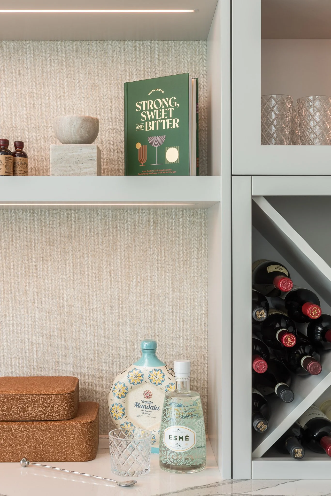 A close-up of a home bar shelf with a green cocktail recipe book titled 'Strong, Sweet and Bitter,' a beige bowl, liquor bottles, a decorative ceramic pitcher, a liquor bottle, a glass, and a brown box on a countertop, with wine bottles stored sidewa