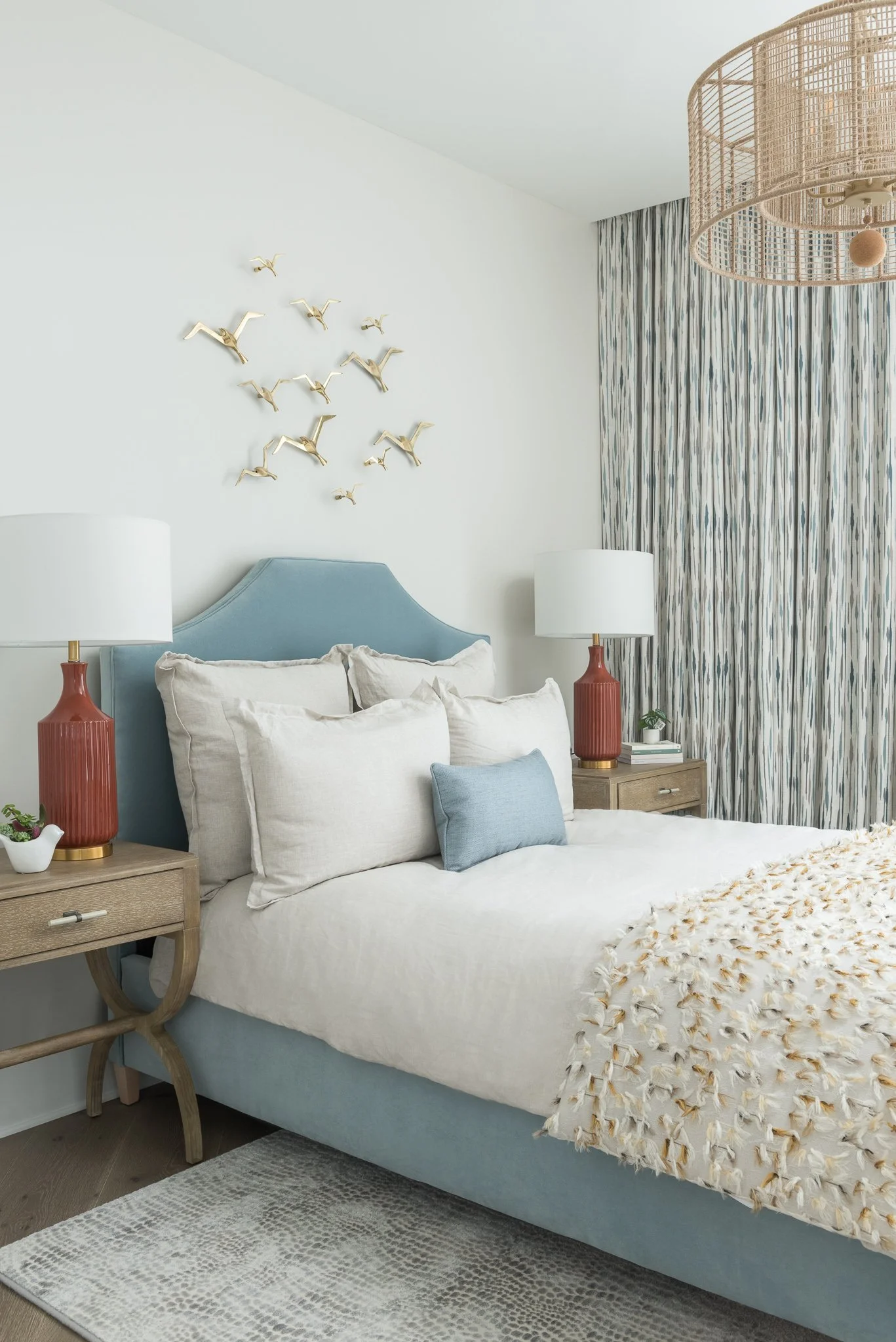 A bedroom with a light blue upholstered bed, cream-colored pillows, and a textured cream blanket. Two wooden nightstands with matching red lamps and small plants. Opposite the bed, a large window with multicolored patterned curtains. Wall decor of go