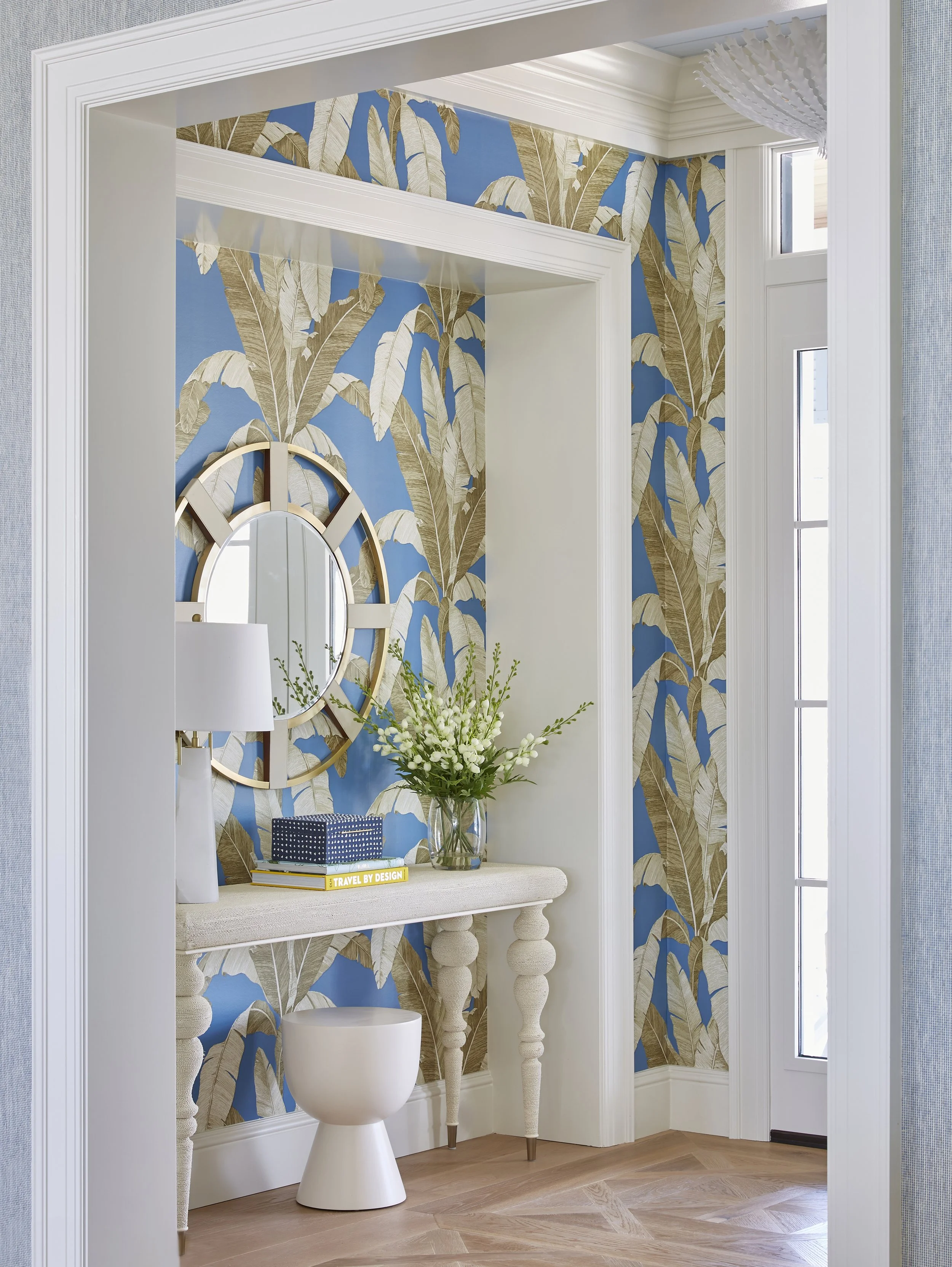 A hallway with blue and gold banana leaf patterned wallpaper, a round mirror with a wooden frame, a white console table decorated with a blue box, books, a white lamp, flowers in a glass vase, and a white stool beneath.