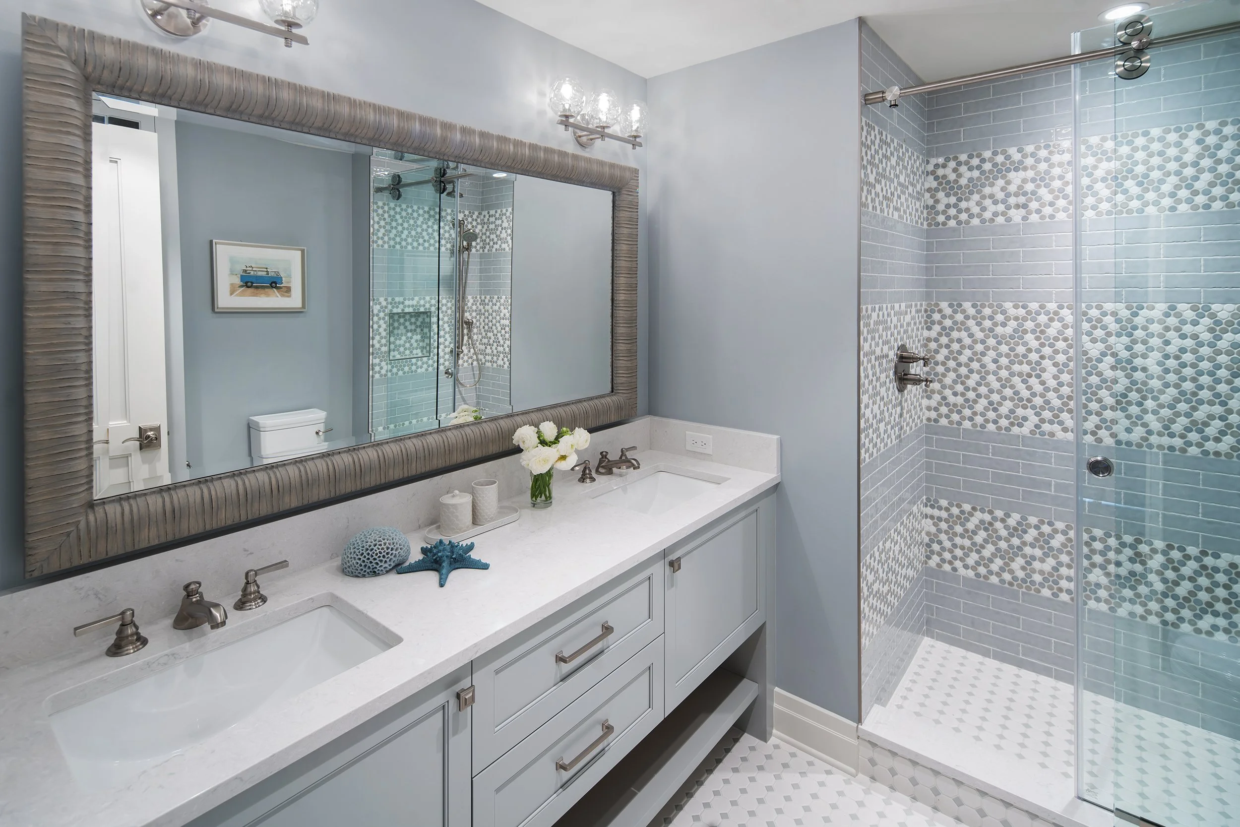 Modern bathroom with light blue walls, large framed mirror, and white vanity with dual sinks. Decor includes flowers and nautical items. Walk-in shower has gray and white patterned tiles and a glass door.
