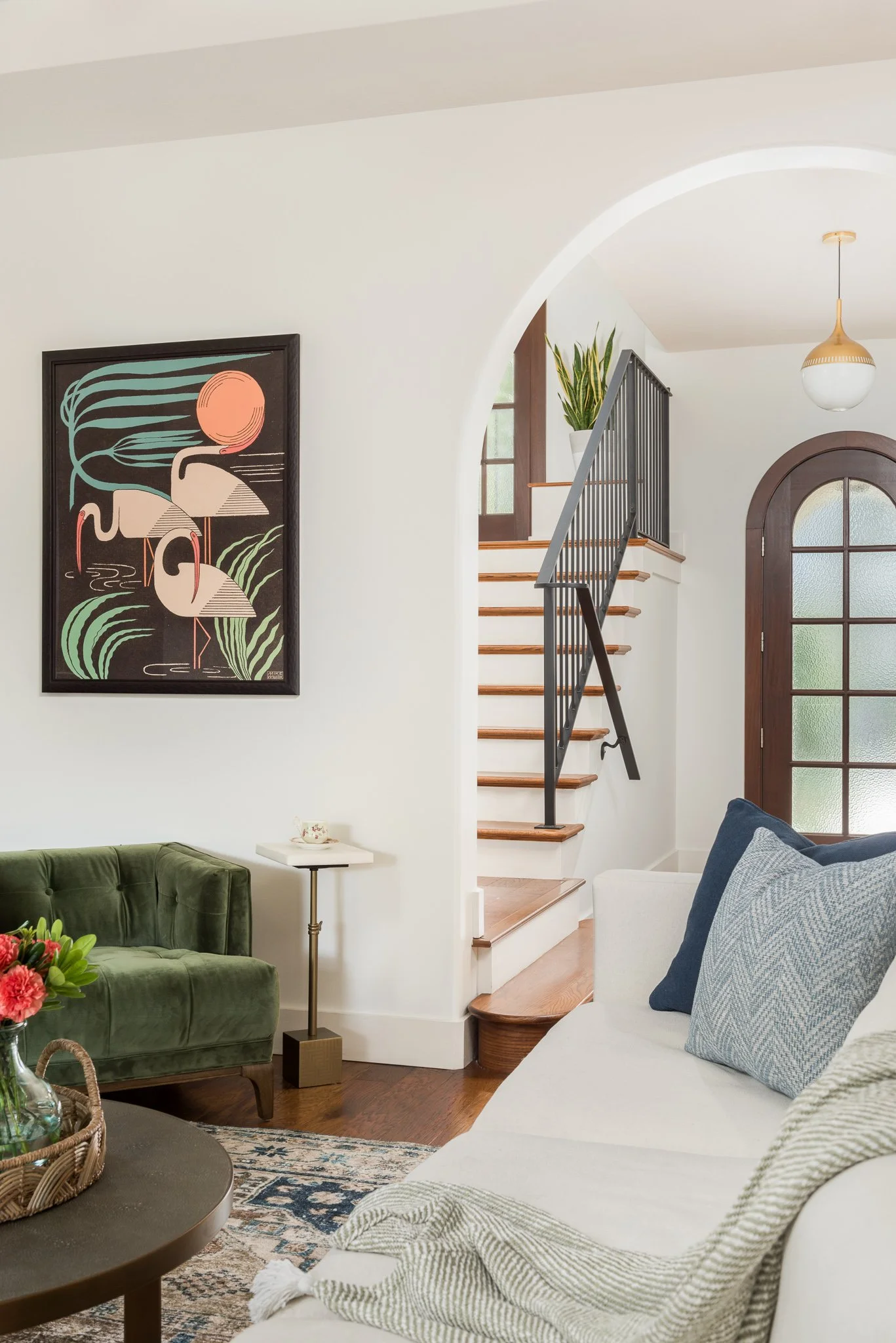 Living room with staircase, framed art of flamingos, green velvet armchair, white sofa with blue cushions, wooden side table, and a white hanging light fixture.