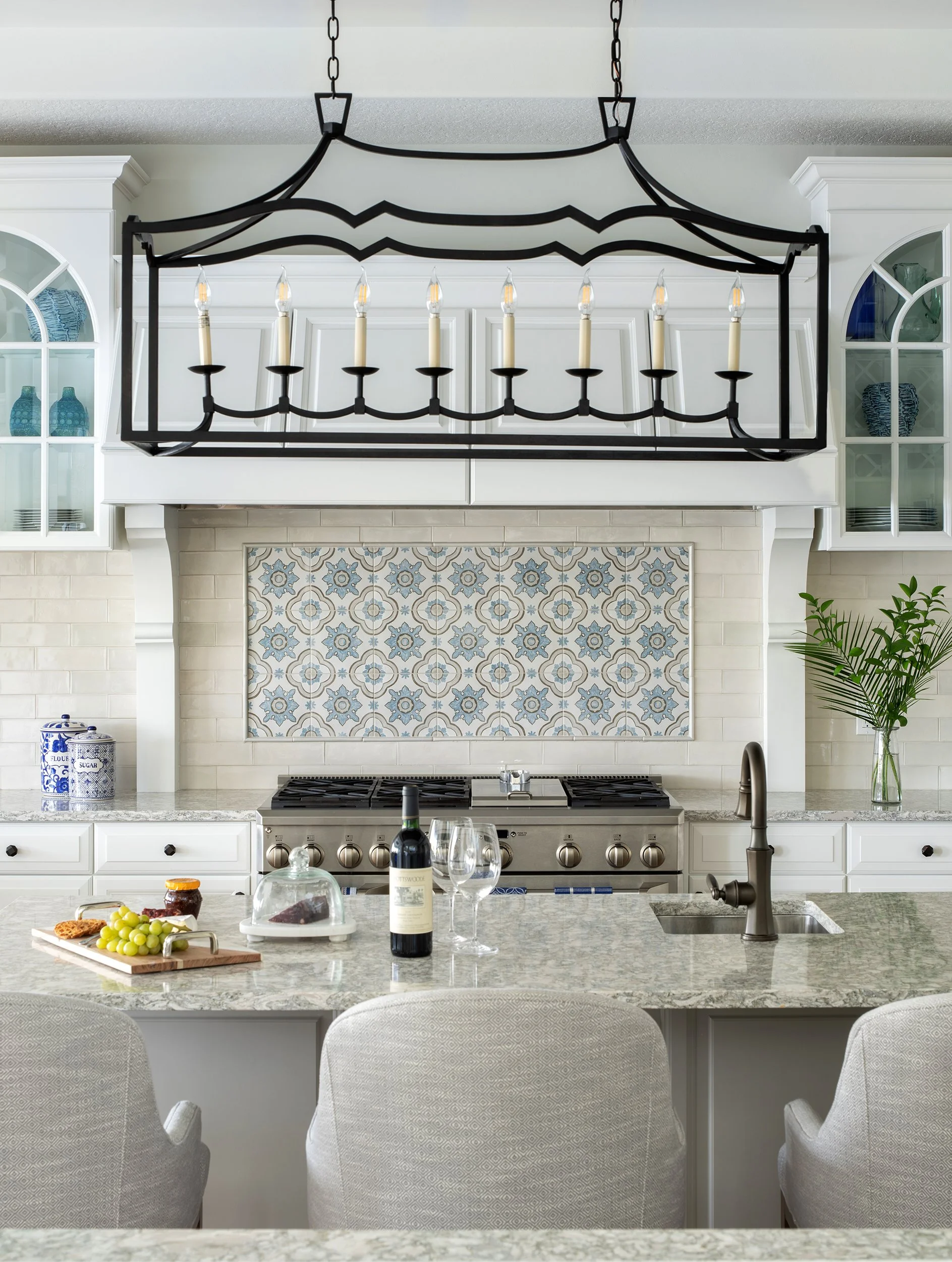 A modern kitchen with white cabinetry, a patterned tile backsplash, and a large black metal chandelier with candle-like lights hanging above the kitchen island.