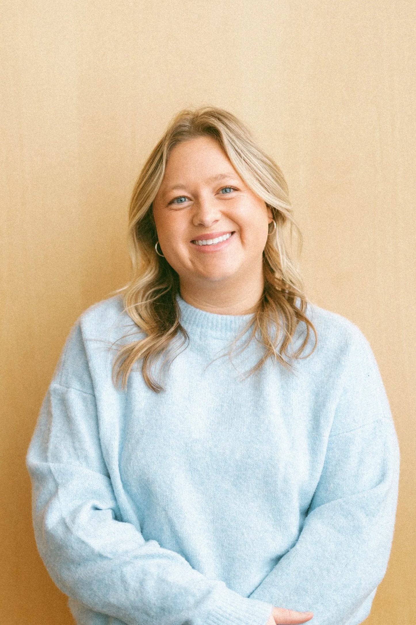 A woman with blonde hair and blue eyes smiling, wearing a light blue sweater, standing against a beige background.
