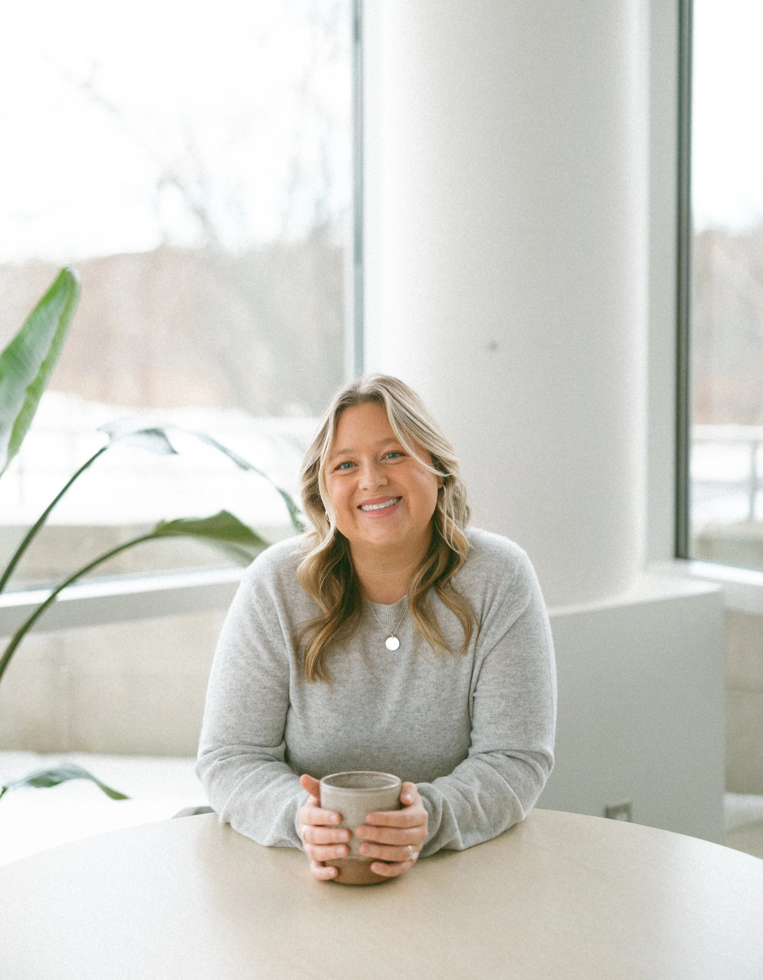 A woman with blonde hair smiling and holding a ceramic mug while sitting at a light-colored table in a bright room with large windows and a plant in the background.