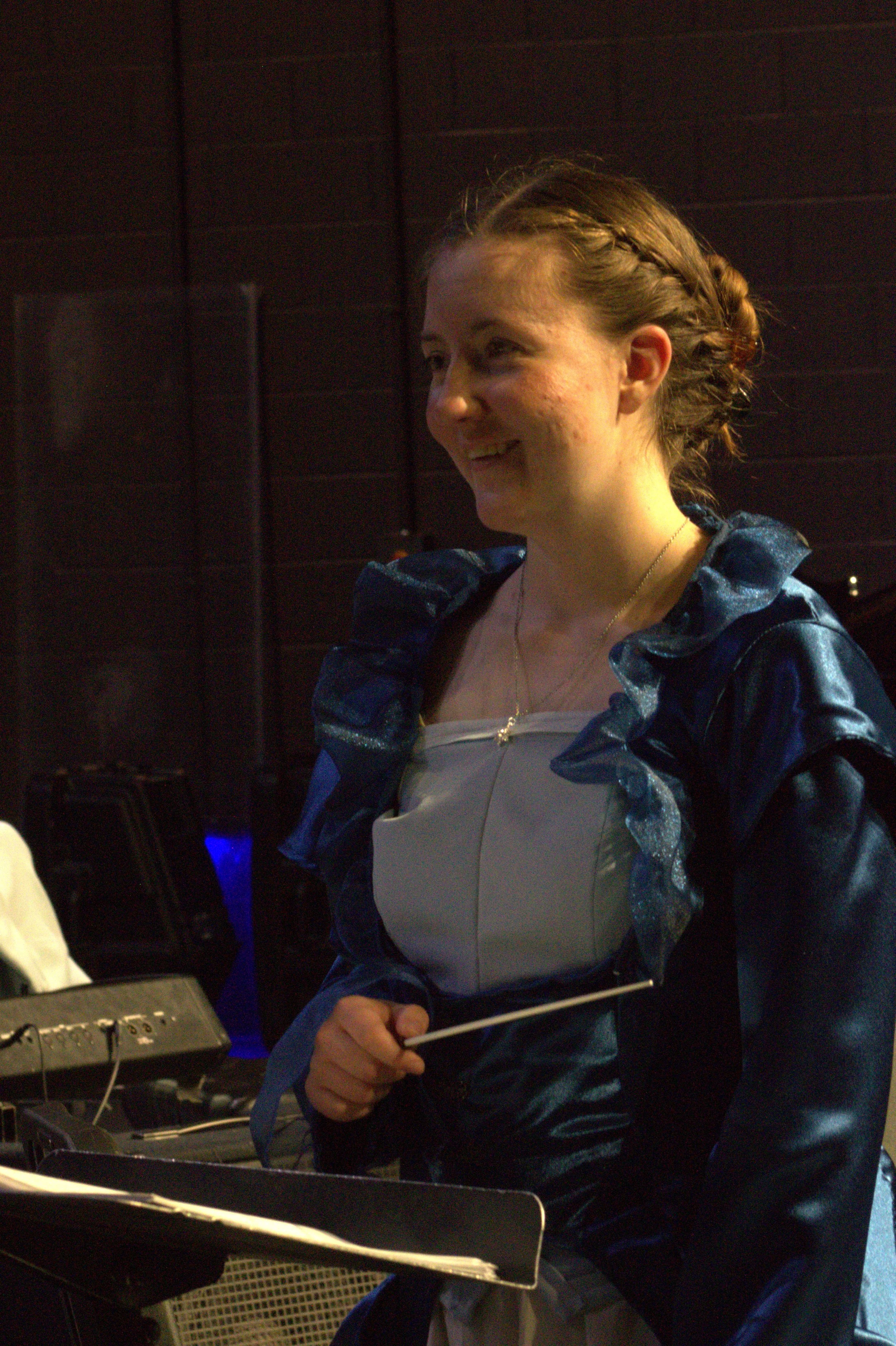A young woman with braided hair smiling, wearing a fancy dress with a shiny, ruffled dark blue jacket, standing near musical equipment.