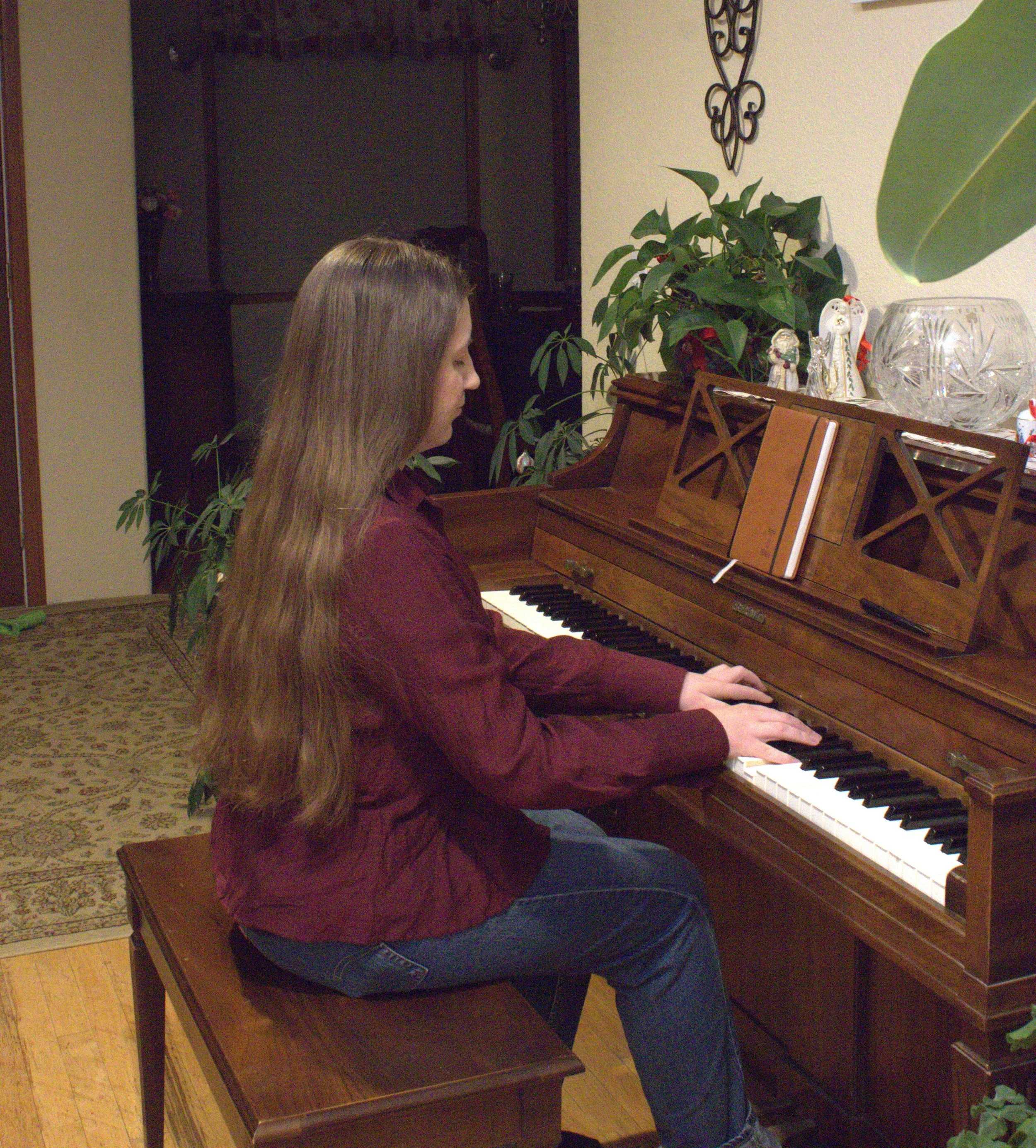 A girl with long brown hair wearing a maroon shirt and blue jeans sitting at a wooden piano with sheet music on the music stand, playing the piano inside a living room with plants and decorative items.