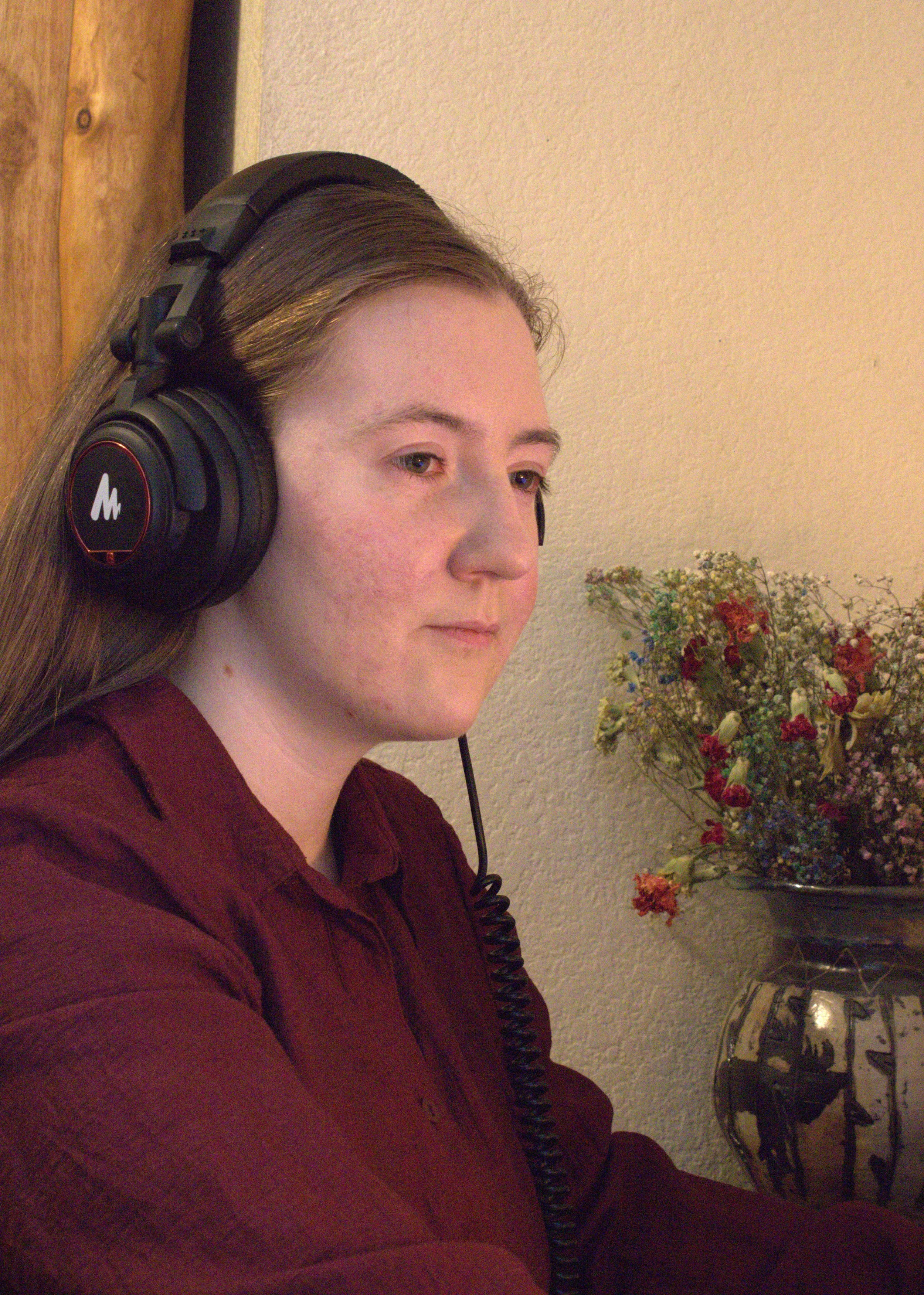 A young woman with long brown hair wearing a maroon shirt and large black headphones sitting indoors near a beige wall and a flower vase.