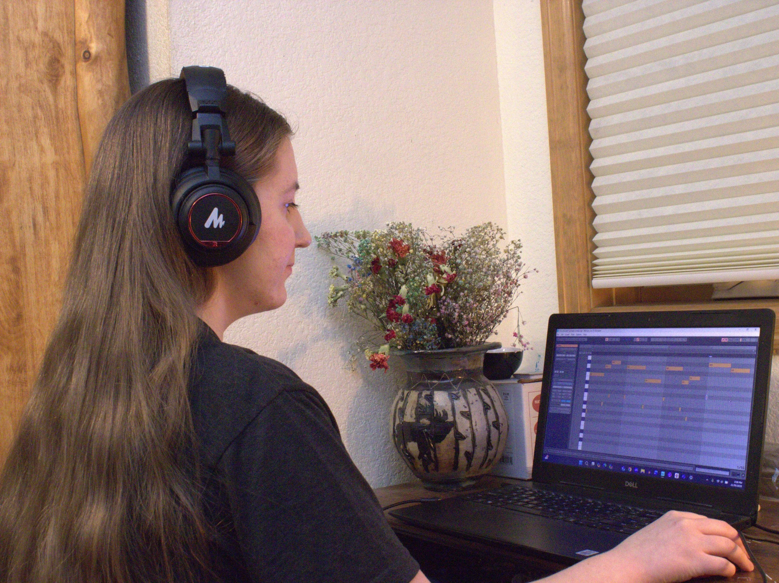 A woman with long brown hair wearing large black headphones sits at a desk working on a music production software on a laptop, with a vase of flowers next to her and a window with closed blinds behind her.