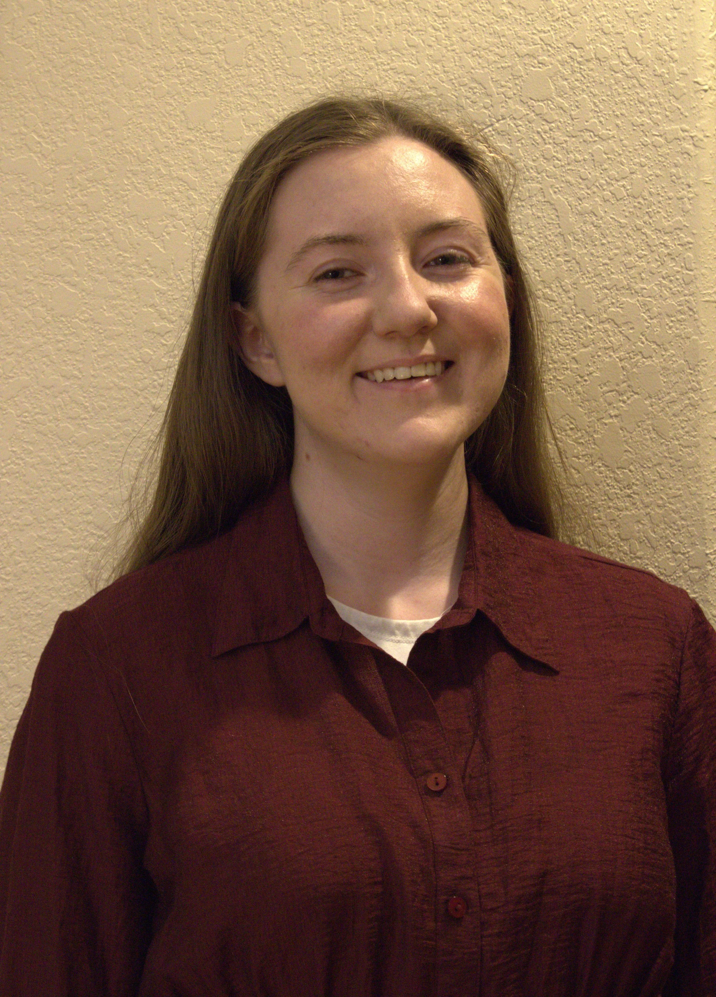 A young woman with long brown hair smiling, standing against a beige textured wall, wearing a maroon button-up shirt.