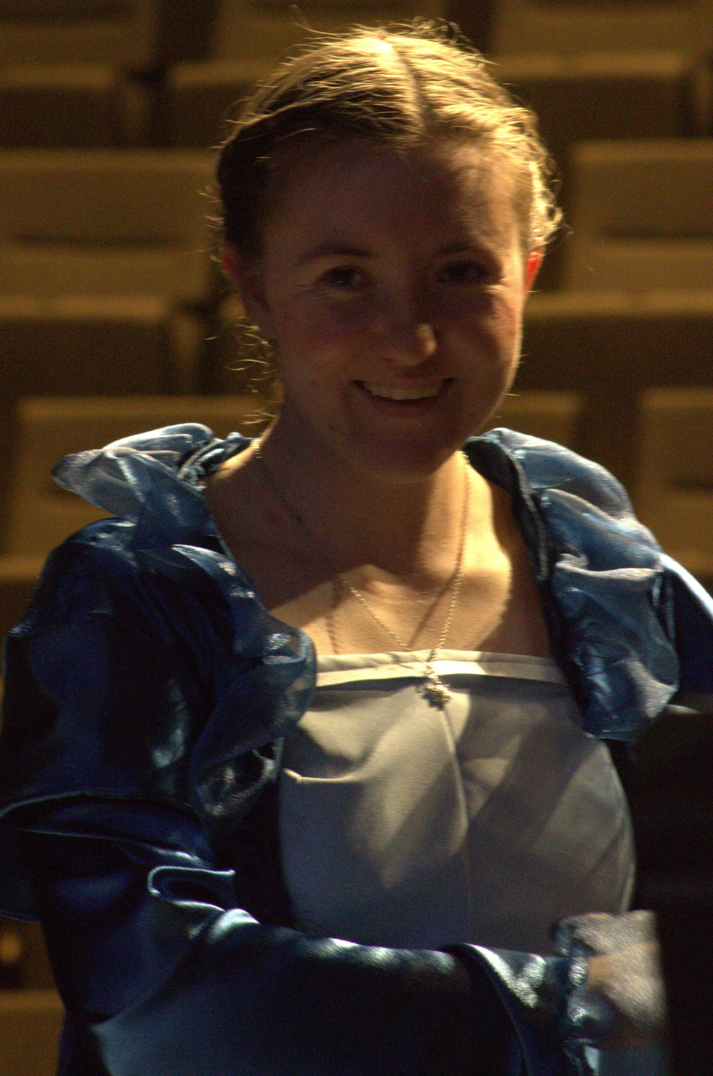 Smiling woman with braided hair wearing a shiny blue jacket and gray top, standing outdoors in front of a stadium or arena seating.