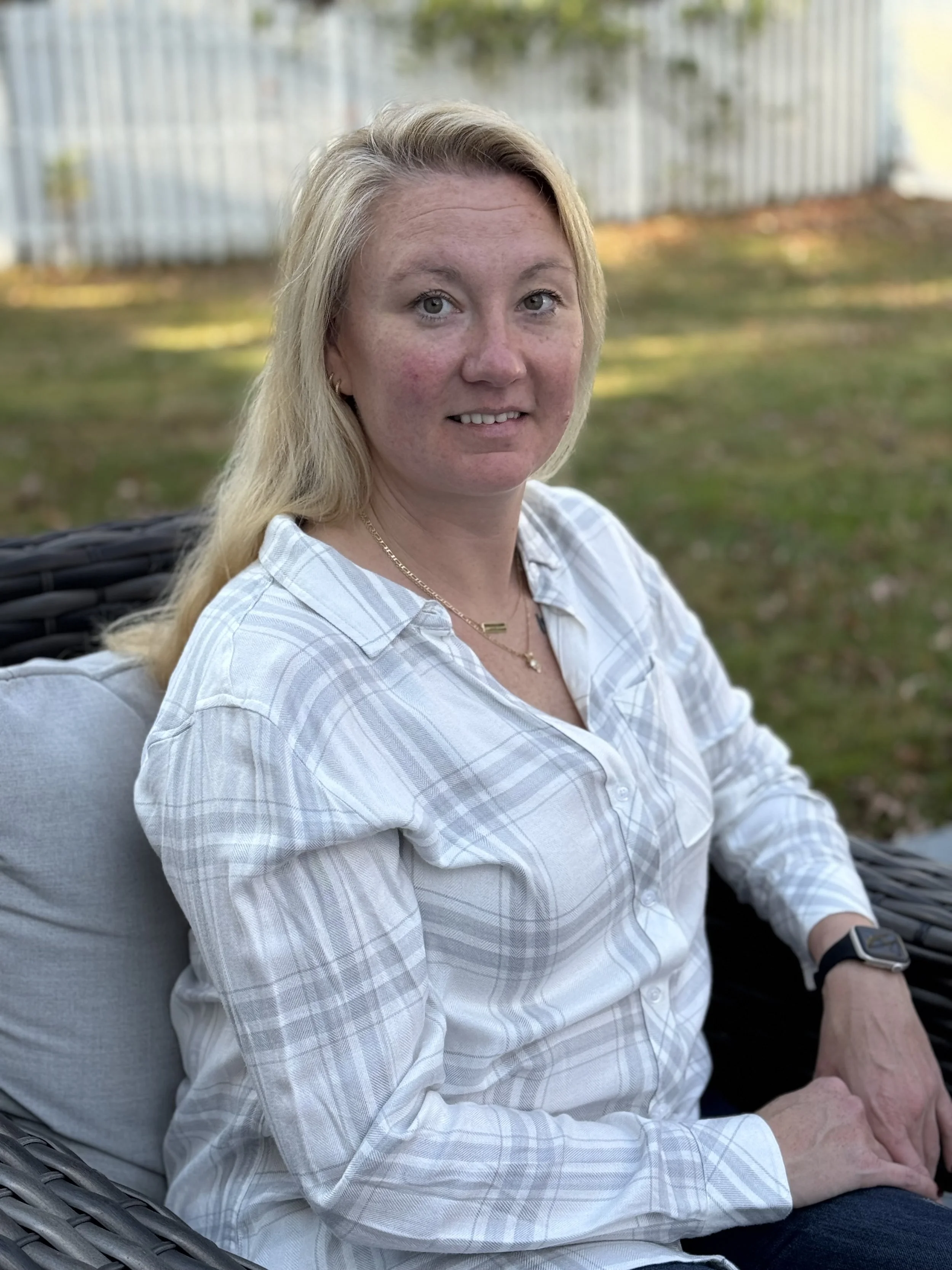 A woman with blonde hair, wearing a white plaid shirt, sitting outdoors on a wicker chair with a cushion, in front of a wooden fence and grassy yard.