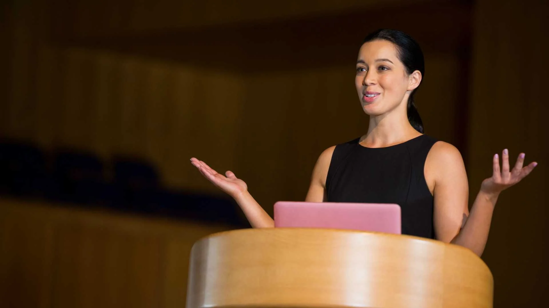 Woman giving a presentation or speech at a podium with a pink laptop in front of her, wearing a sleeveless black dress, inside a room with dark background.