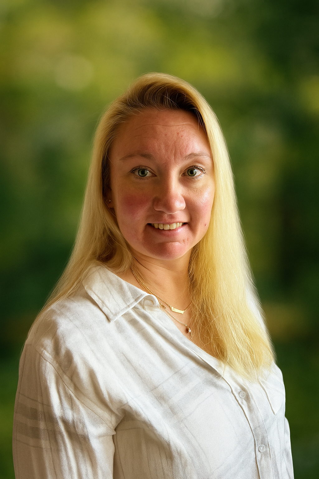 A woman with long blonde hair wearing a white button-up shirt, smiling slightly, against a blurred green outdoor background.
