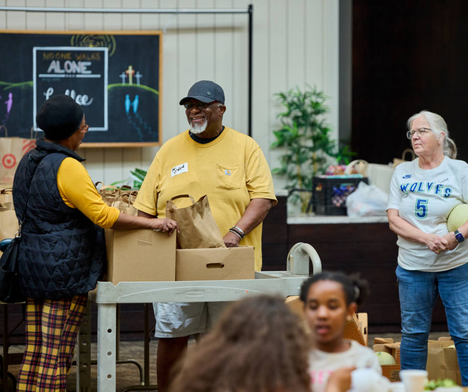 People donating food or supplies at a charity event, with a man in a yellow shirt smiling, and a woman in a white sports jersey standing nearby.