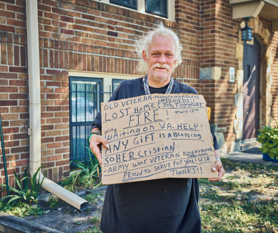 An elderly man with white curly hair and a mustache standing outside in front of a brick building, holding a cardboard sign with handwritten message.