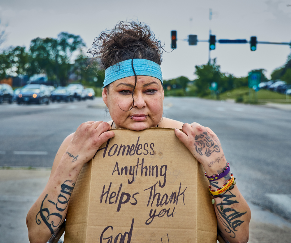 A woman with tattoos, wearing a blue headband, holding a cardboard sign that says 'Homeless Anything Helps Thank you God' on a city street with traffic lights and cars in the background.