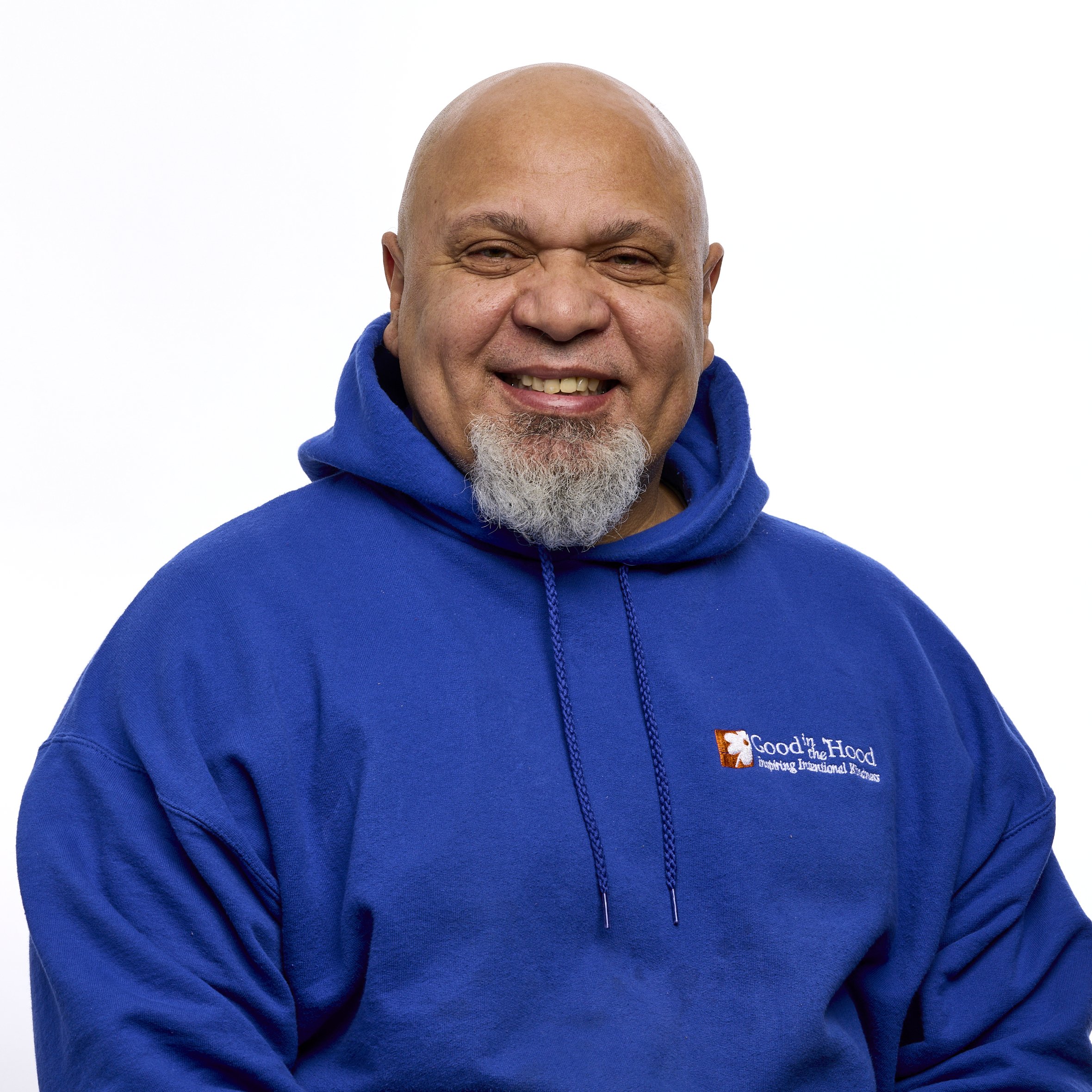A smiling bald man with a beard wearing a blue hoodie with 'Good in the Hood' logo, standing against a plain white background.