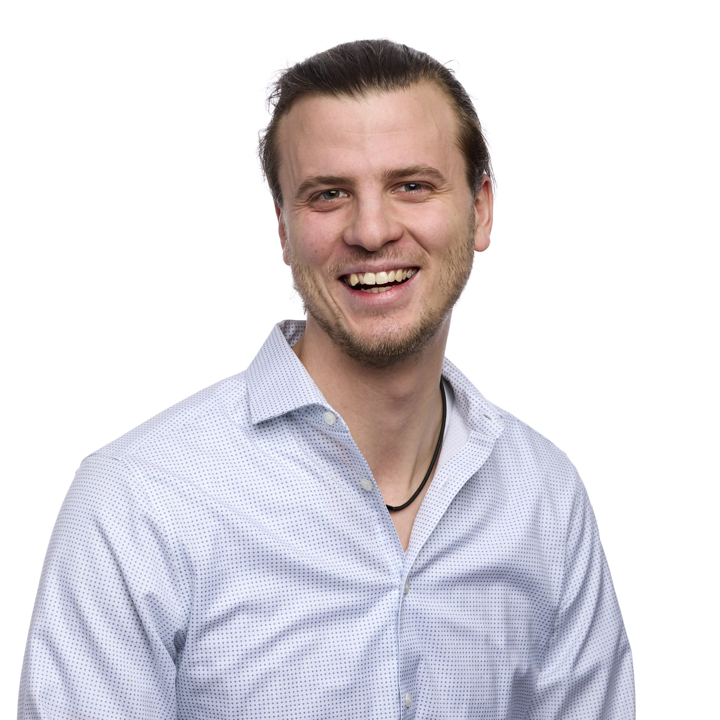 A smiling young man with light skin, brown hair, and a short beard, wearing a light blue collared shirt with small white dots and a black necklace, posed against a white background.