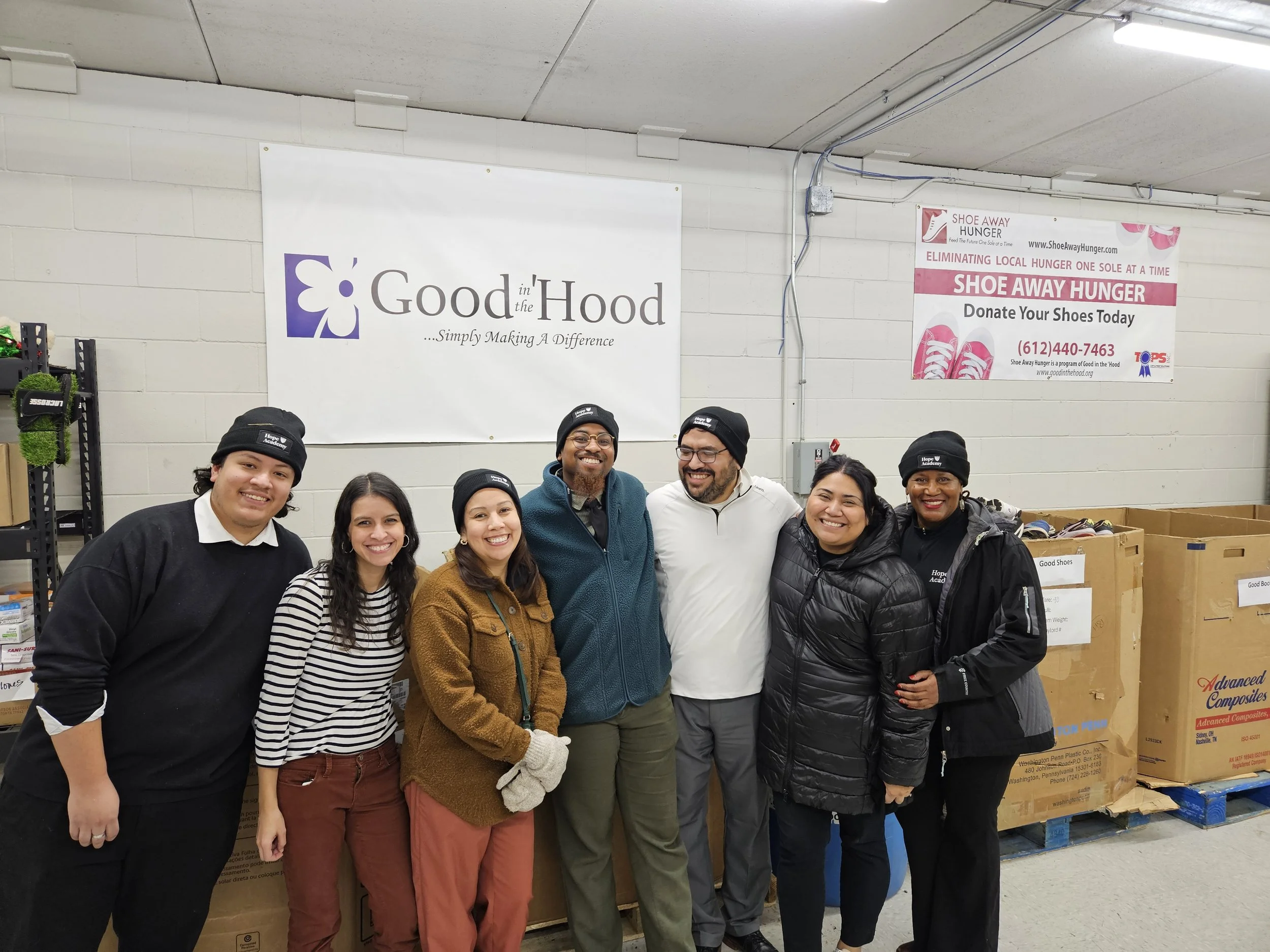 Group of seven people standing together smiling in a warehouse, with boxes and signs behind them that read 'Good in the Hood' and 'Shoe Away Hunger'.