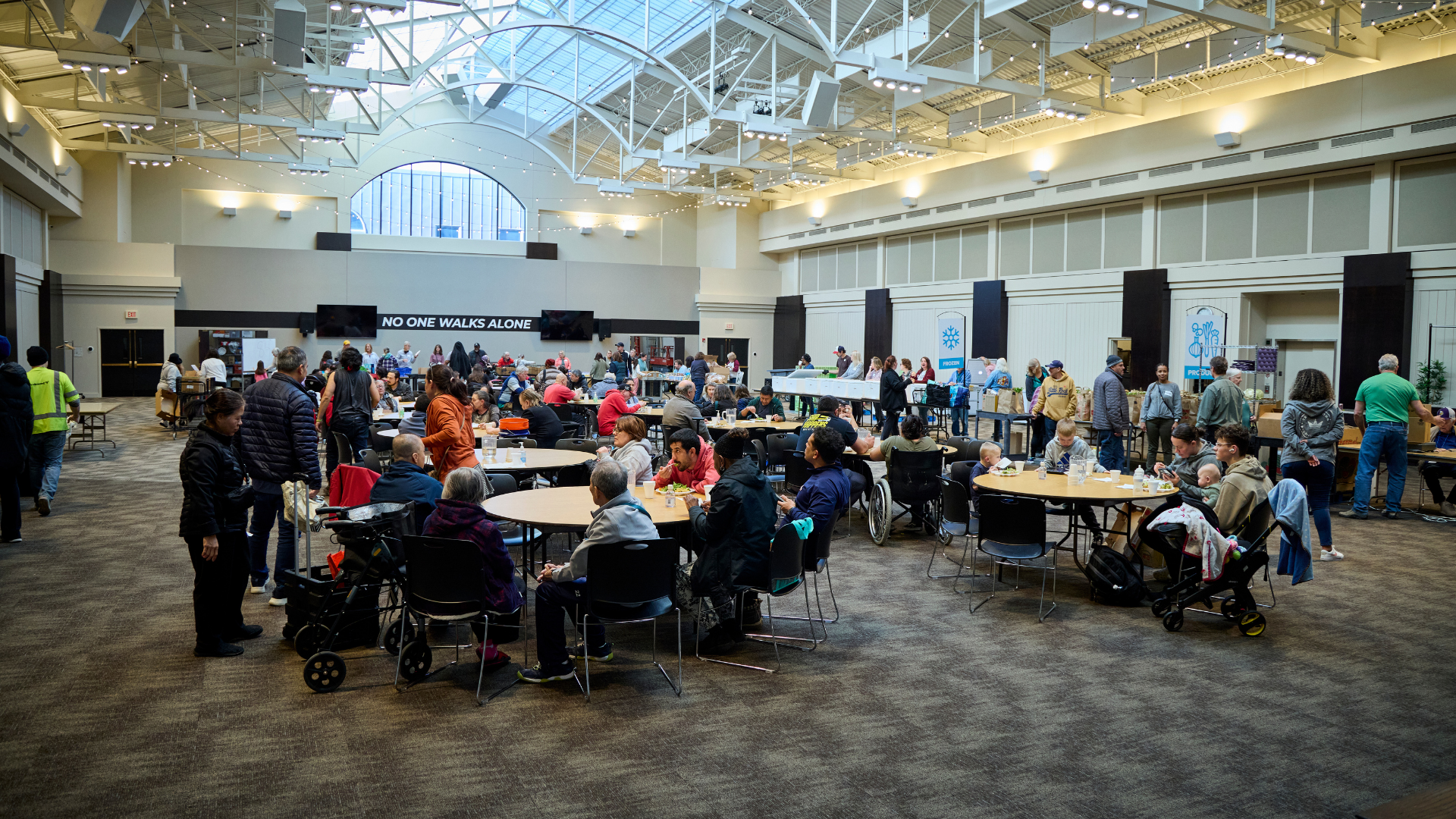People sitting and eating at tables in a large indoor space, with some standing and walking around, in a banquet or community event setting.