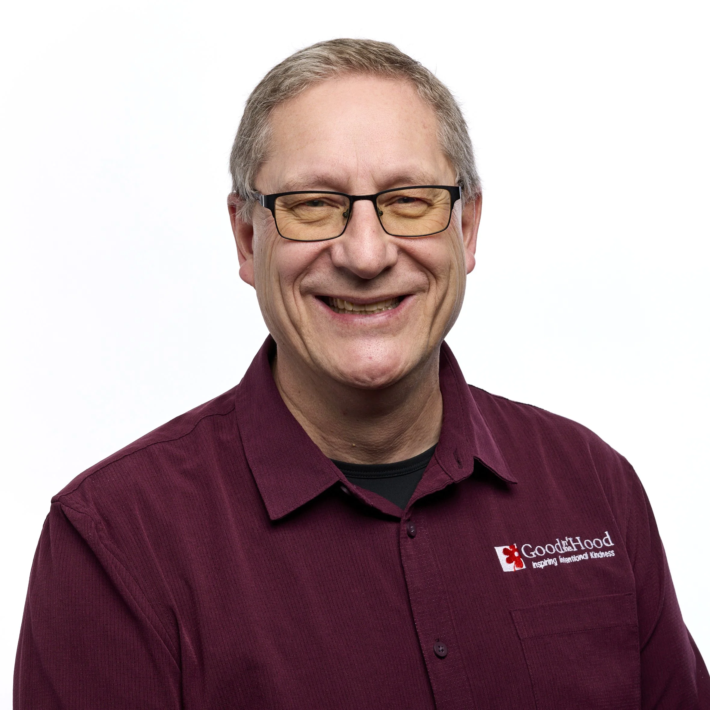 Smiling middle-aged man with gray hair and glasses, wearing a maroon shirt with a logo that reads 'Good Samaritan' and a black undershirt, standing against a white background.