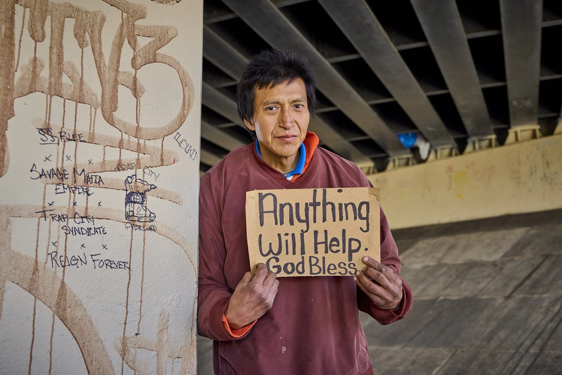 A man holding a cardboard sign that reads 'Anything Will Help. God Bless' standing next to a mural with a map of San Francisco neighborhoods.