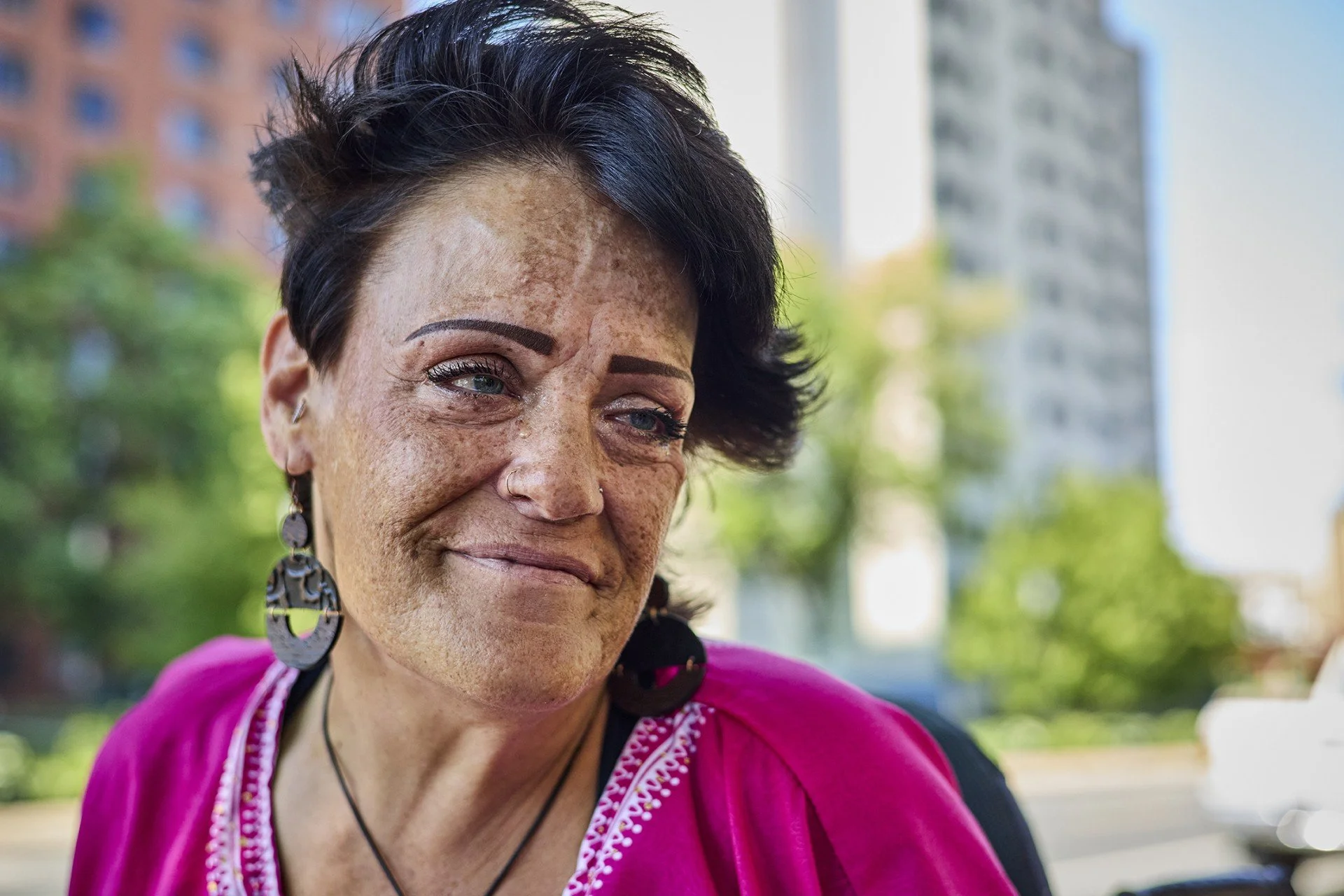 A woman with short dark hair, blue eyes, and freckles wearing earrings and a pink top, smiling outdoors with buildings and trees in the background.
